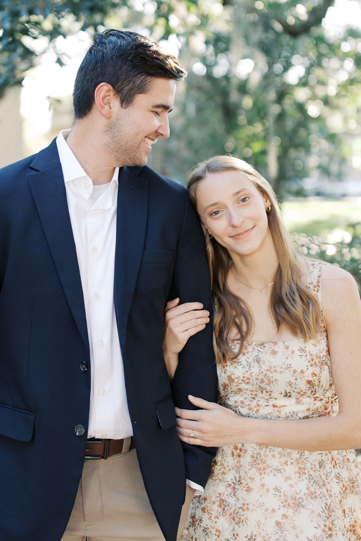 Couple in navy suit and floral dress embracing outdoors in romantic engagement photo.