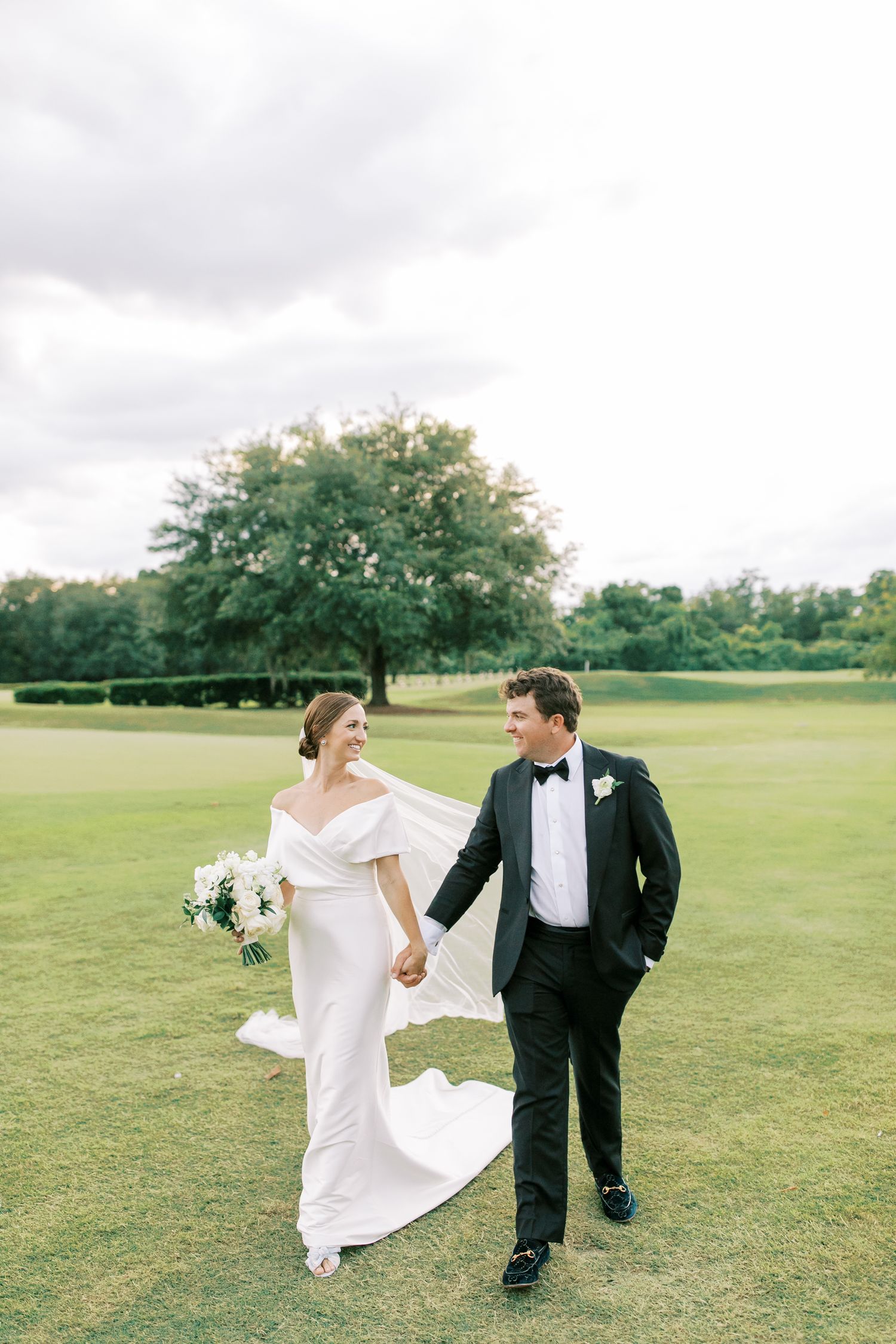 Wedding couple walks hand in hand across lush green lawn with elegant attire and white floral bouquet.