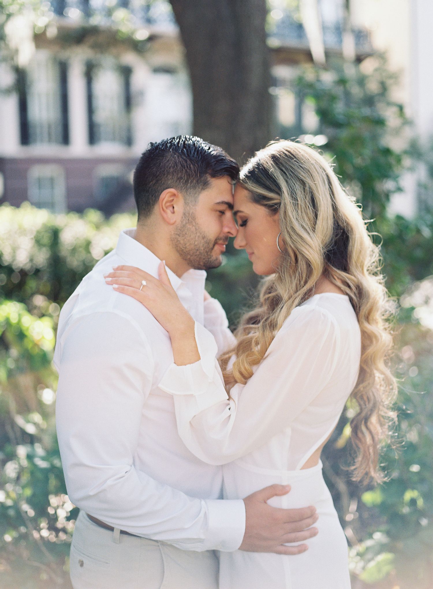 Intimate portrait of couple embracing in white outfits against garden backdrop.