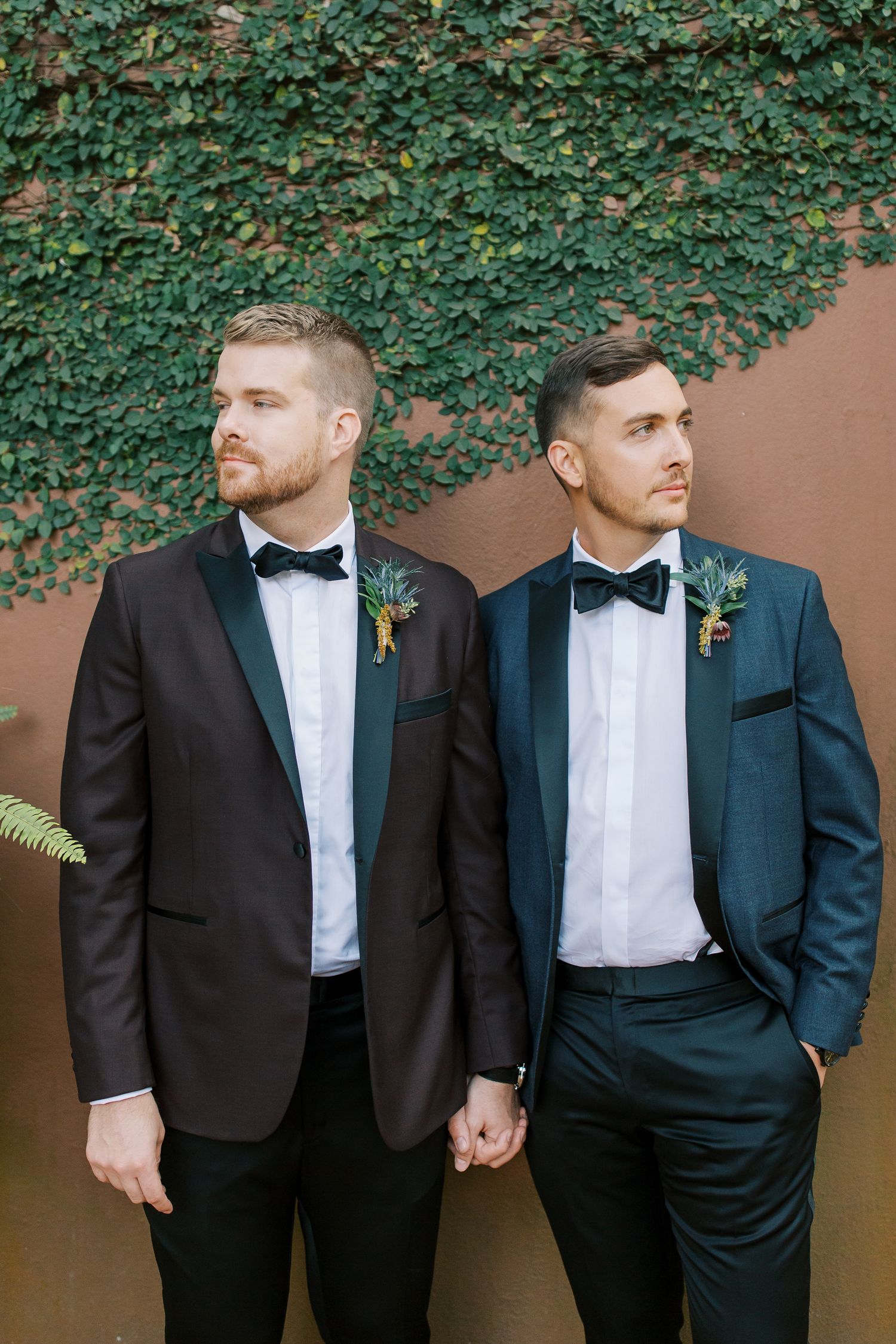 Two grooms in matching black tuxedos and bow ties hold hands while posing against an ivy-covered wall in downtown Savannah.