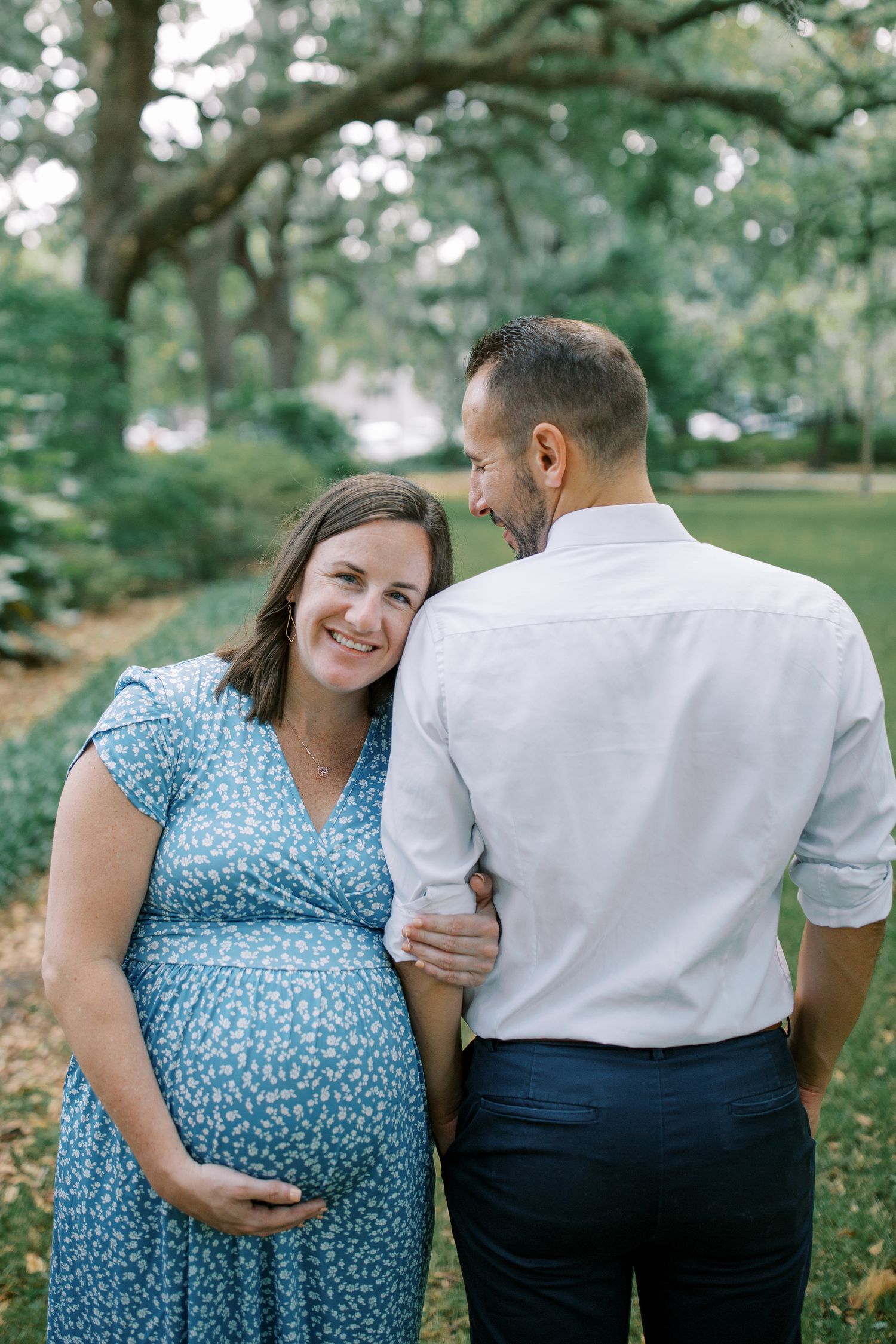 Intimate outdoor maternity photos of a couple in blue dress and white shirt against a leafy backdrop.