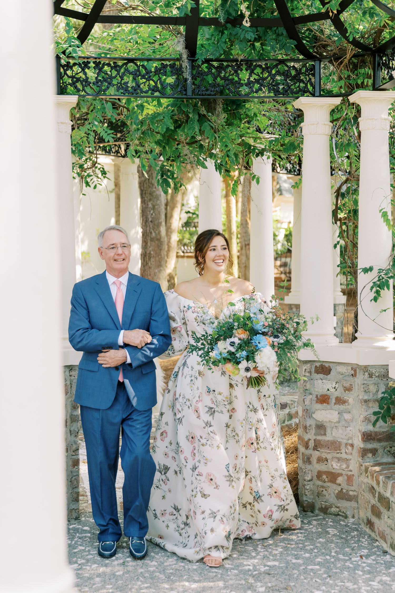 A couple stands together under a covered walkway with white columns and climbing vines.