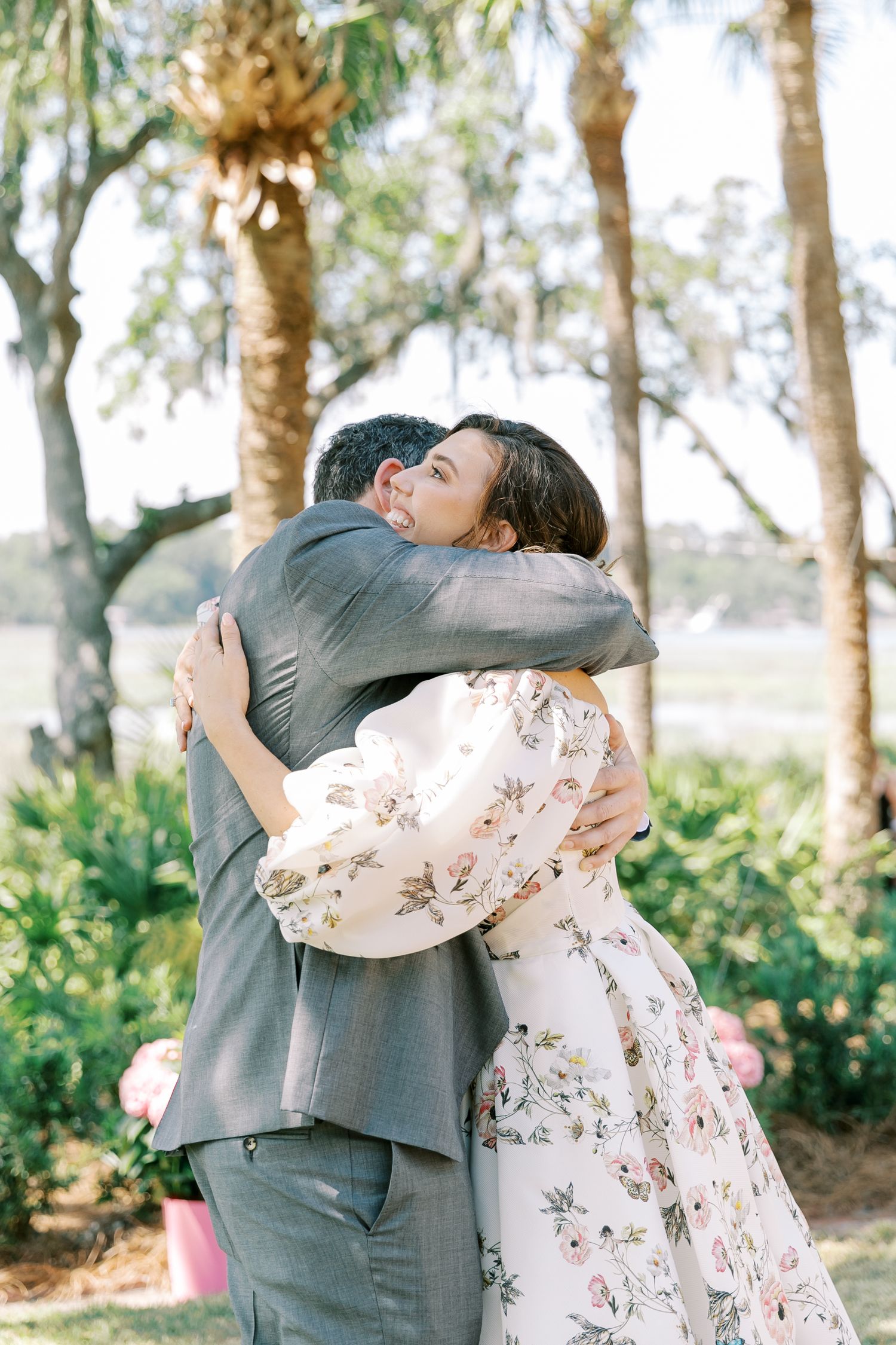 Couple in floral dress and gray suit embrace joyfully in an outdoor garden setting with palm trees.