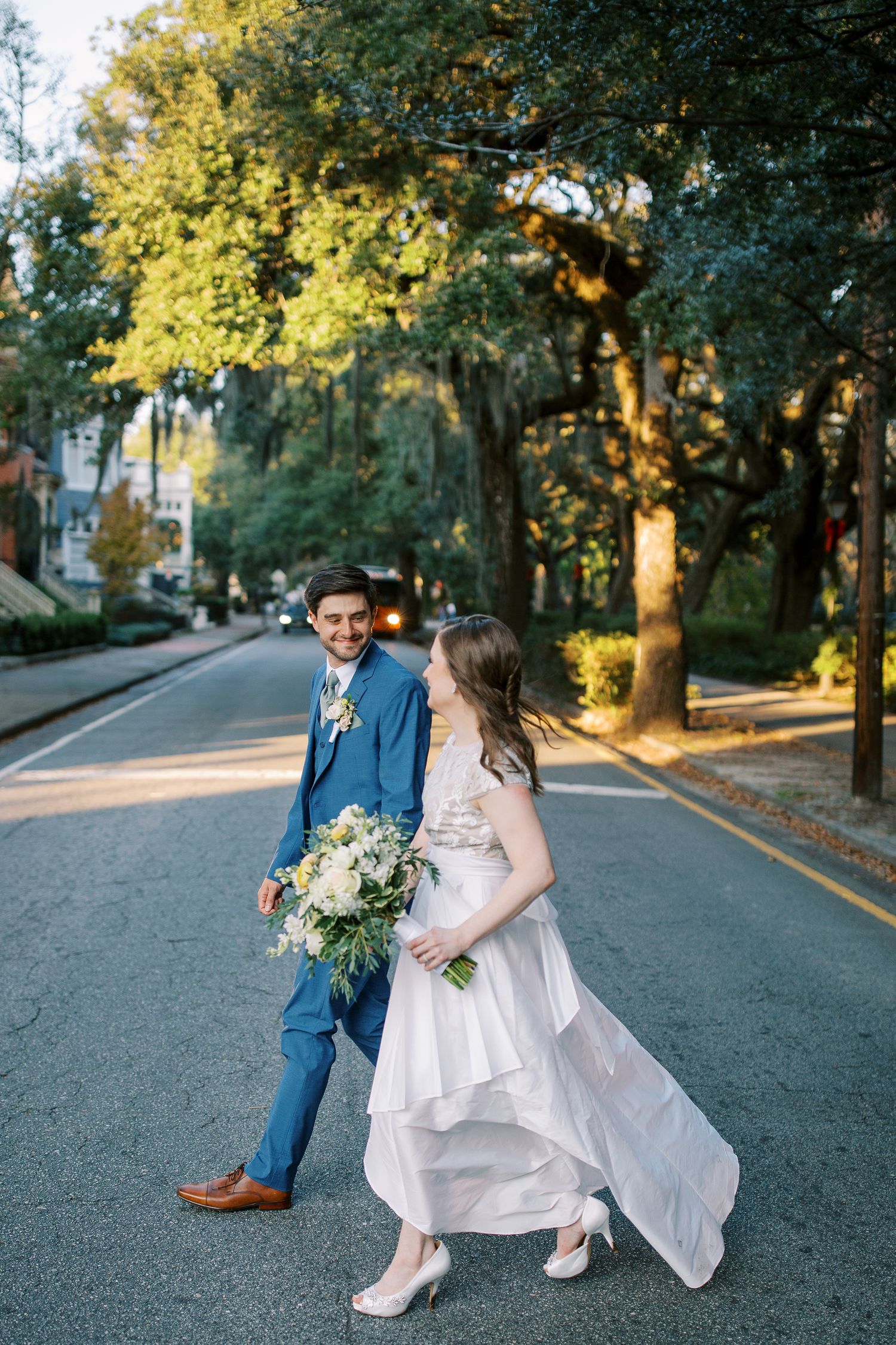 A couple walks down a tree-lined street in wedding attire, with the bride's dress flowing in the breeze.