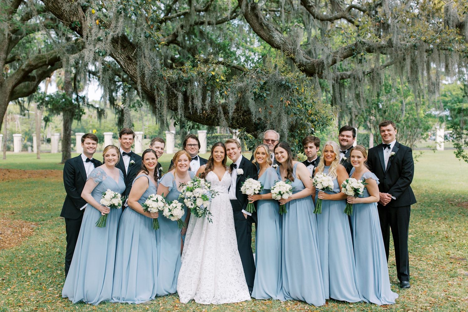 Wedding party in powder blue dresses and dark suits pose together outdoors under oak trees.