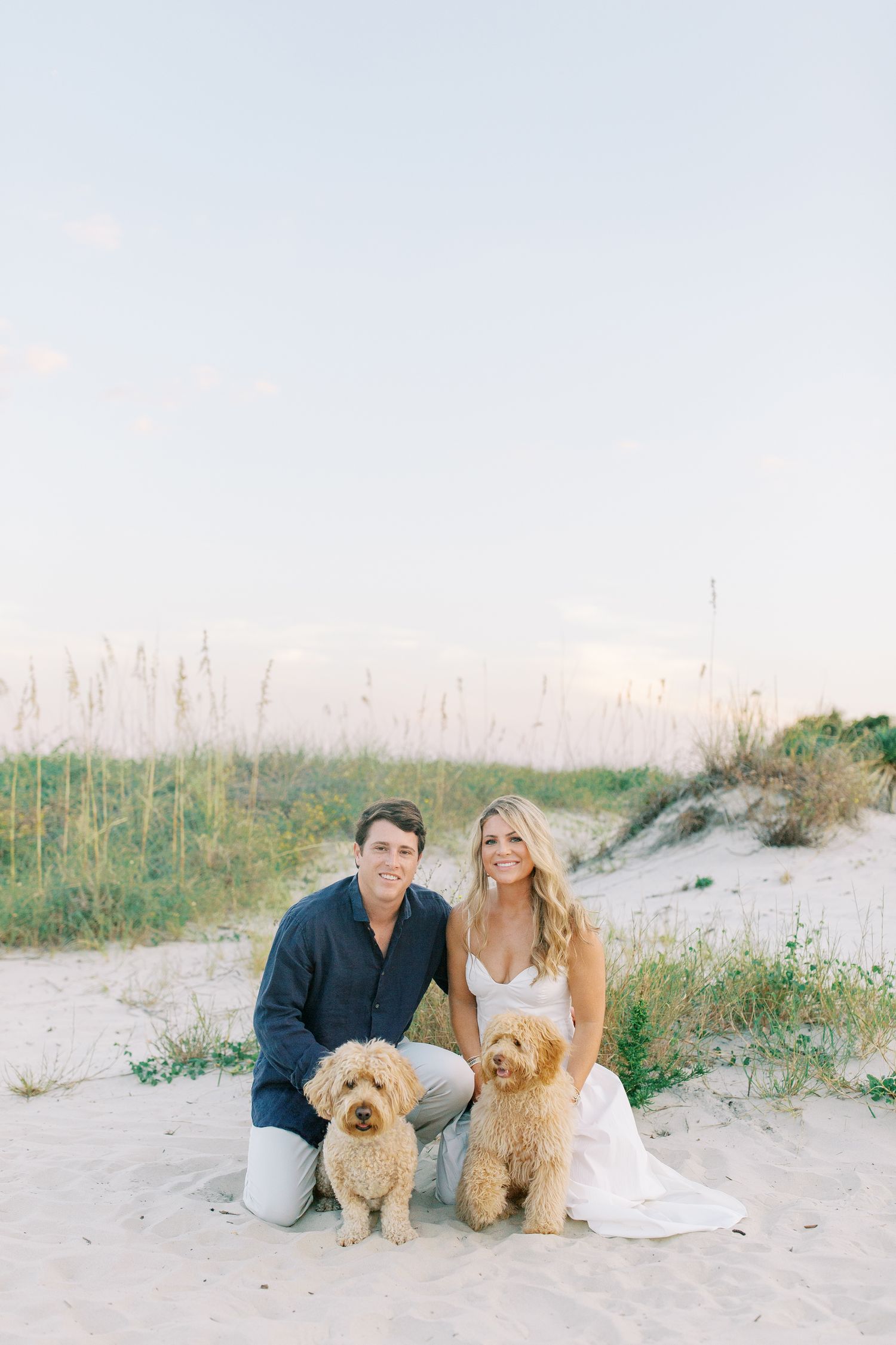 A couple sits with their two goldendoodle dogs on a sandy beach with sea oats in the background during sunset in St. Simons, GA