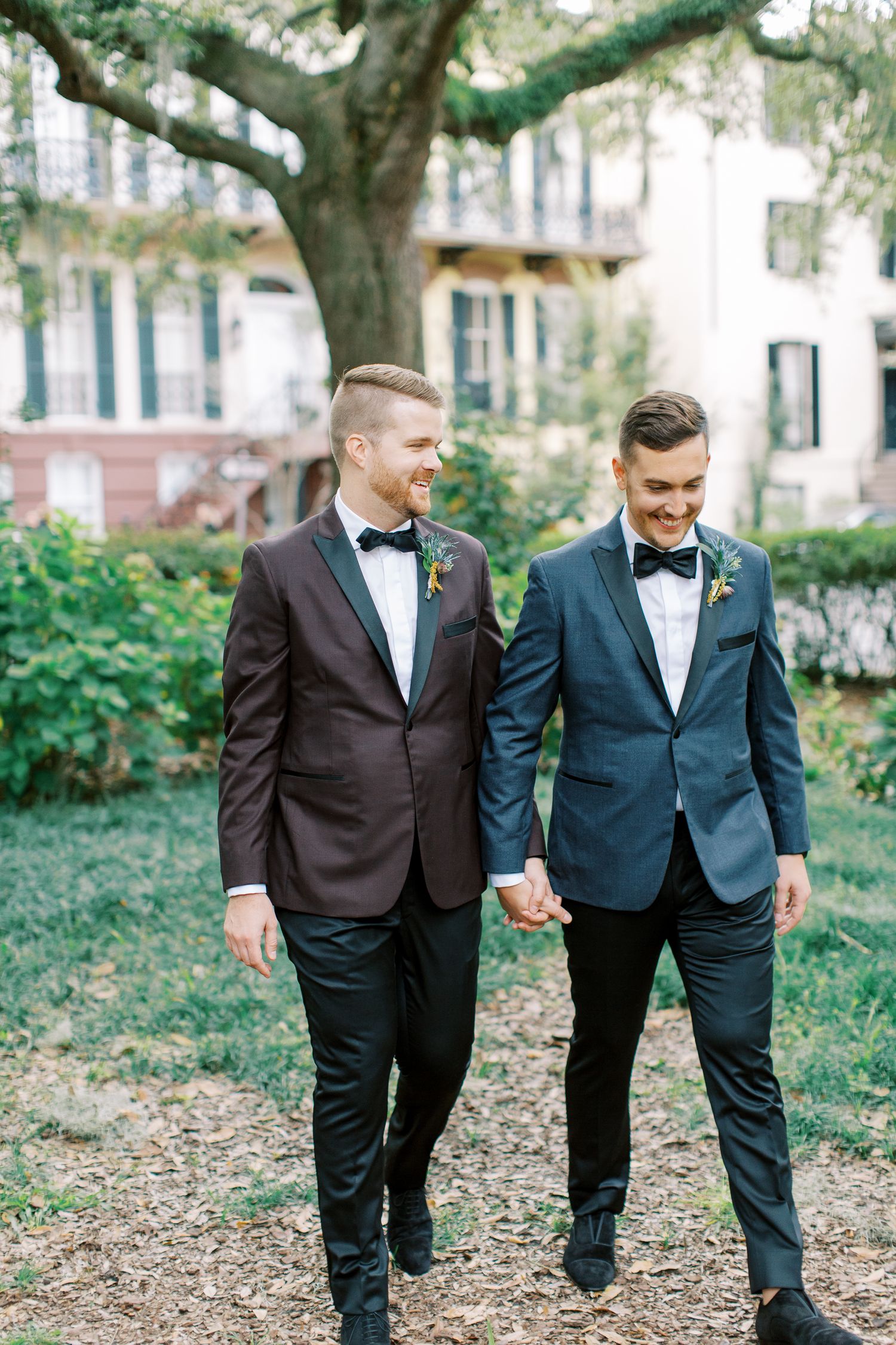 Two grooms in tuxedos holding hands while walking through a garden setting with historic Savannah buildings in background.