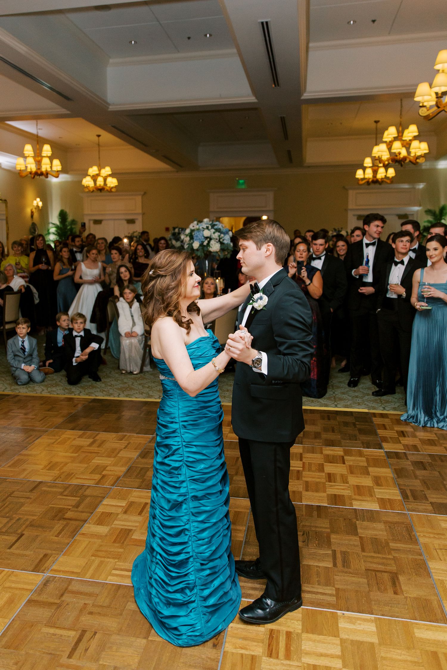 Couple dancing in formal attire on parquet floor with guests watching in background.