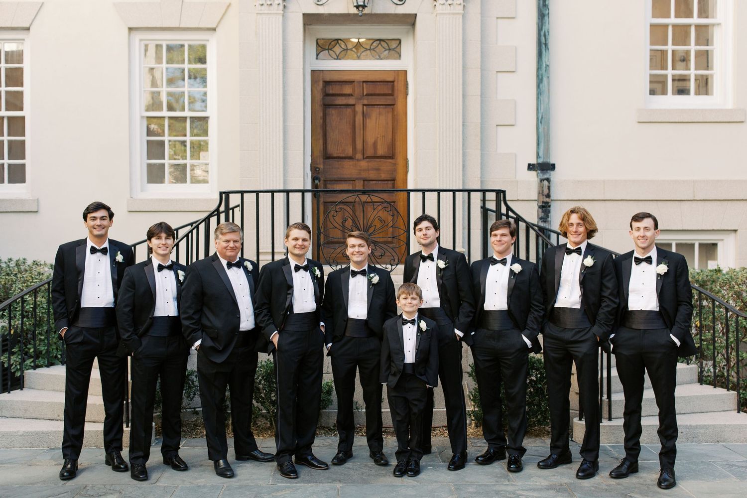 A panoramic shot of groomsmen lined up against a white building exterior in black tuxedos.