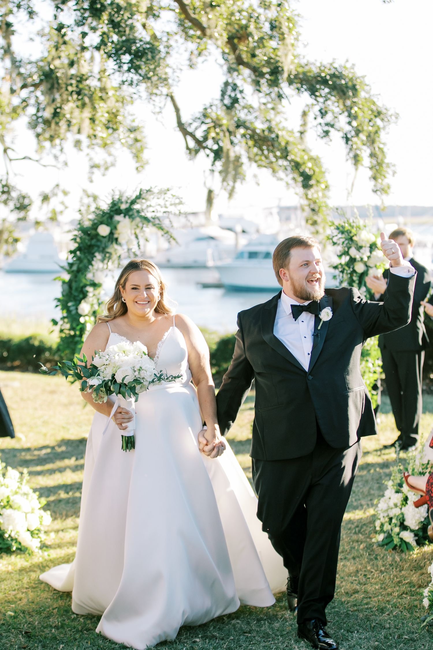 A joyful wedding couple walking outdoors under Spanish moss trees with a floral arch and water view.