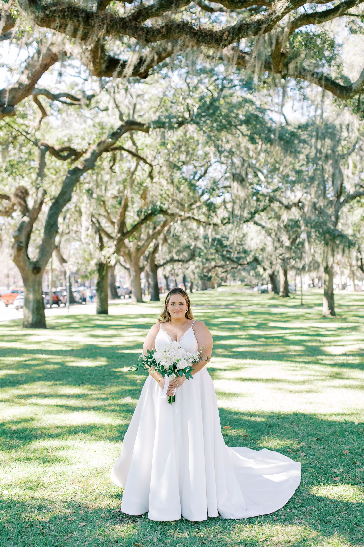 A bride in a classic white ballgown poses under Spanish moss-draped trees in a park setting.