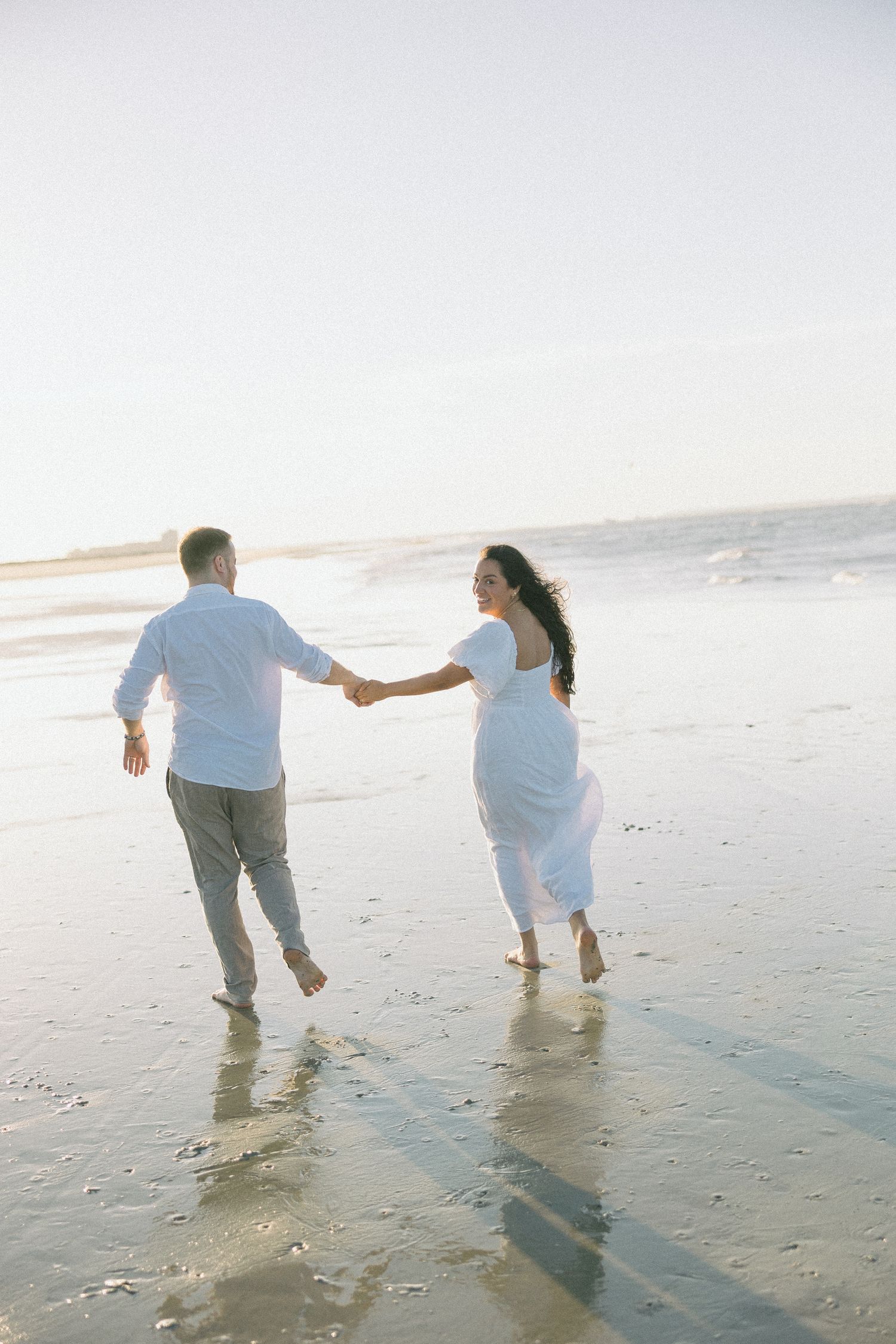 A couple holding hands and walking together on a misty beach during sunset.