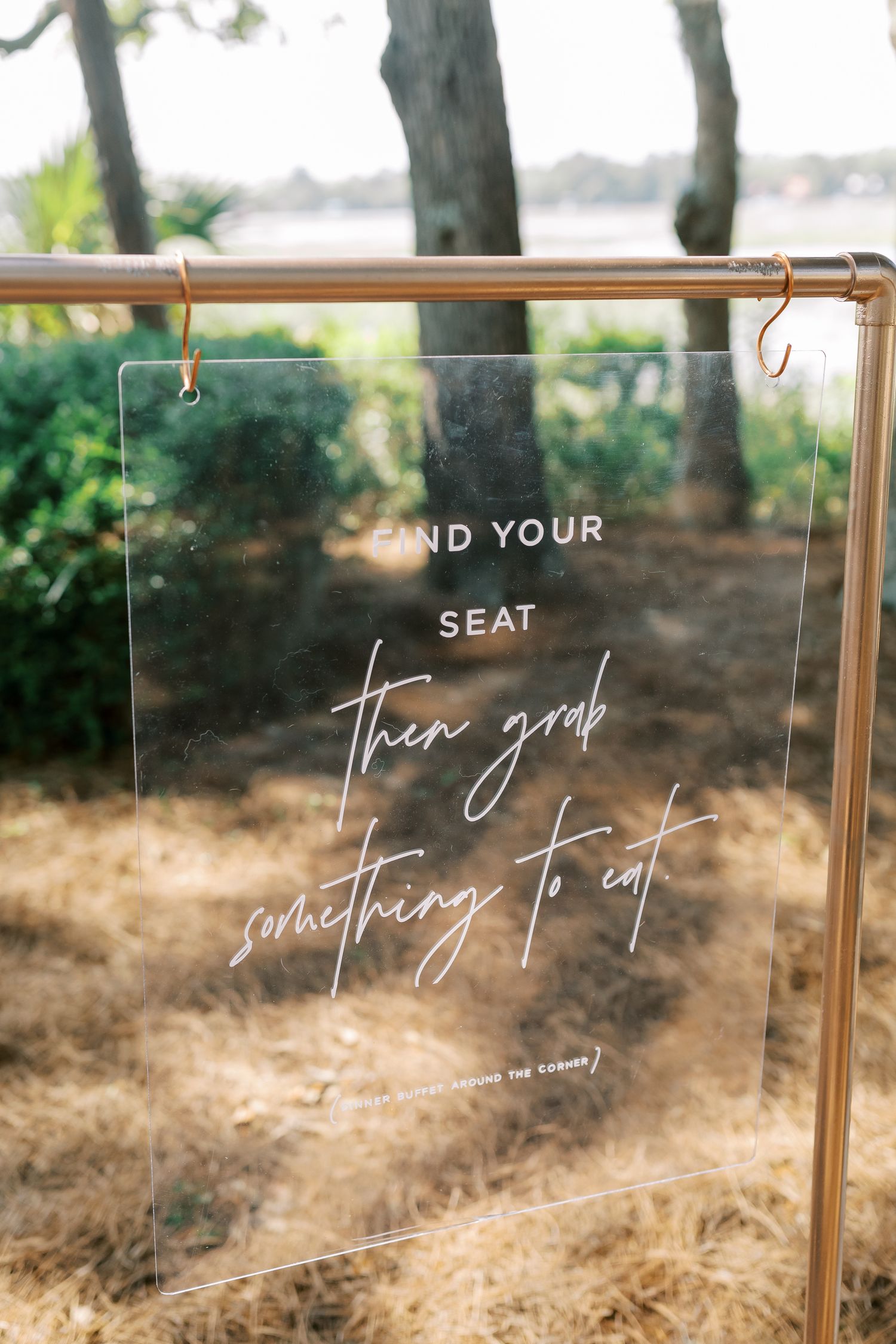 Clear acrylic wedding sign reading 'Find your seat then grab something to eat' hanging on copper rod.