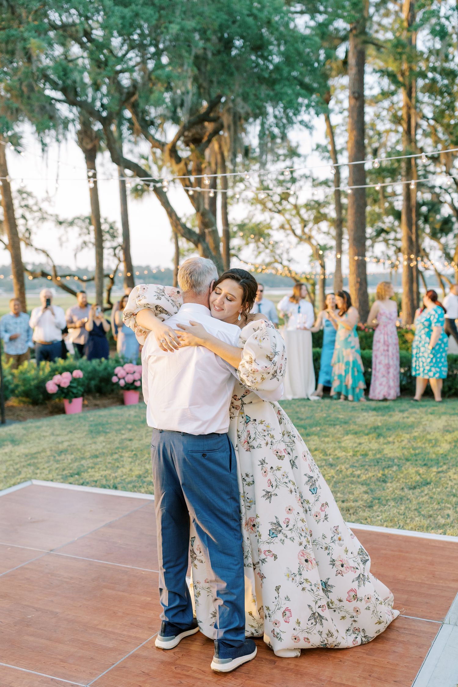 Newlyweds perform their first dance on a tiled patio under trees at sunset.