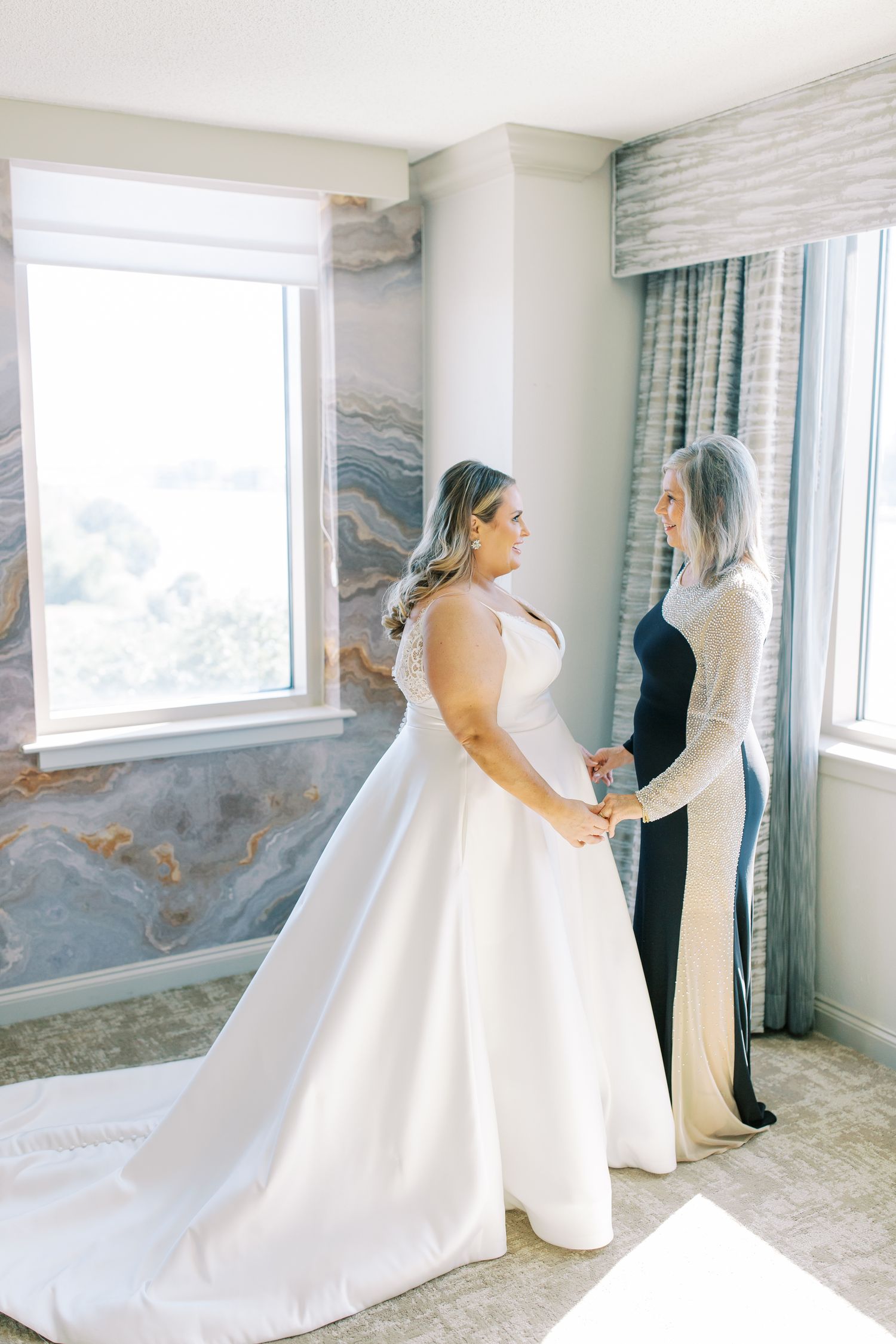Two people in a bright room with large windows share an intimate moment before a wedding ceremony.