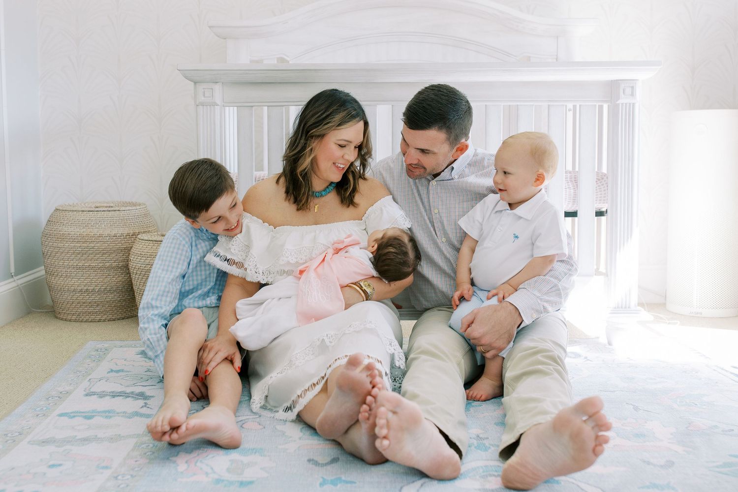 A family relaxes together on a white bed while wearing neutral colored clothes in a bright sunlit room.