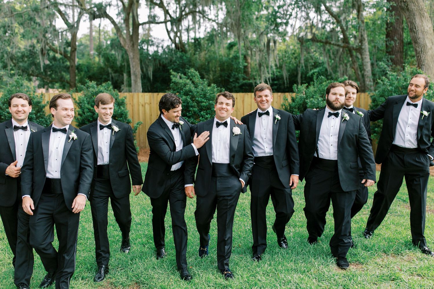 Groomsmen in black tuxedos pose casually on lawn in front of trees.