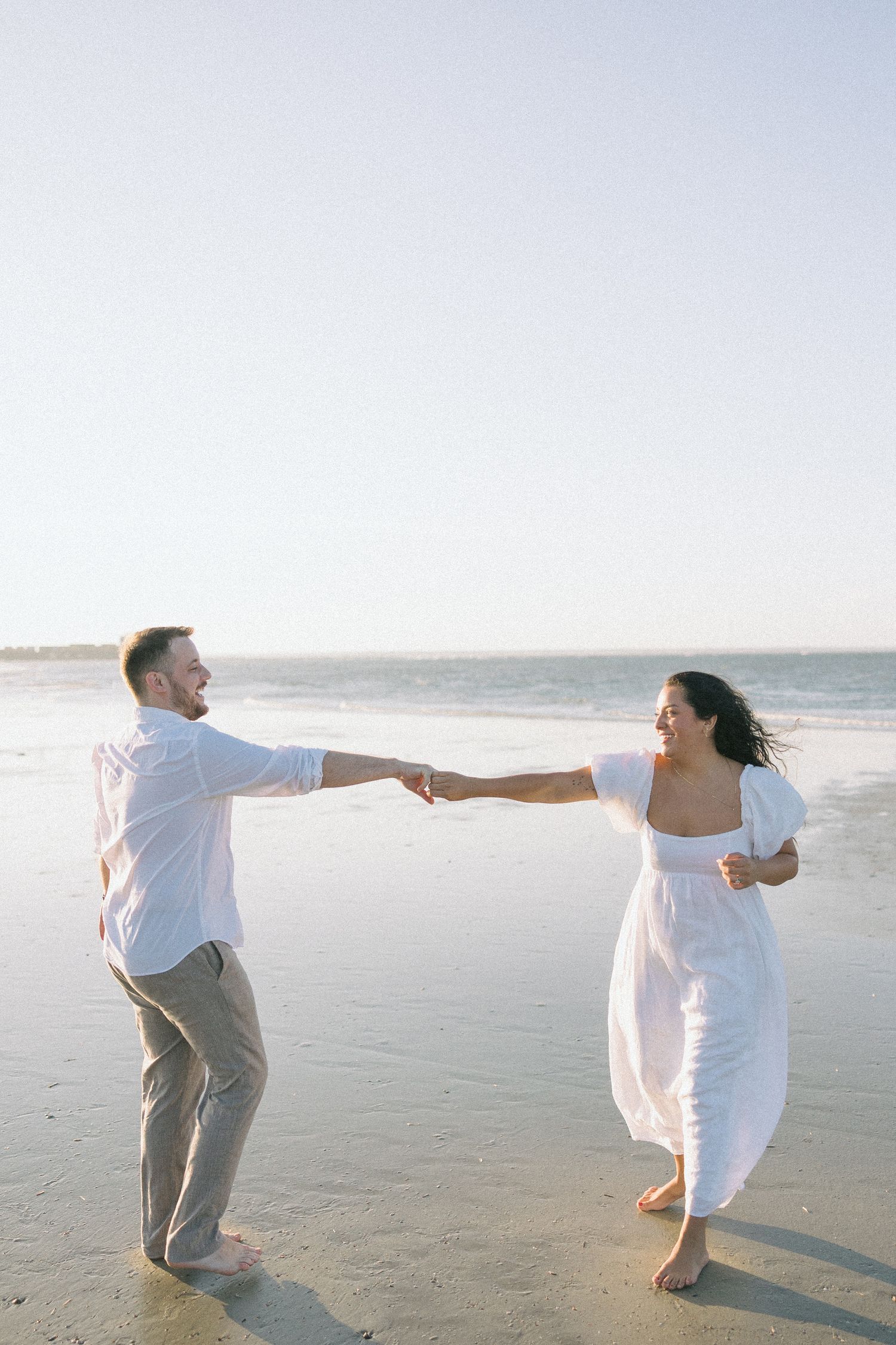 A couple in white clothing dances together on a sandy beach at sunset.