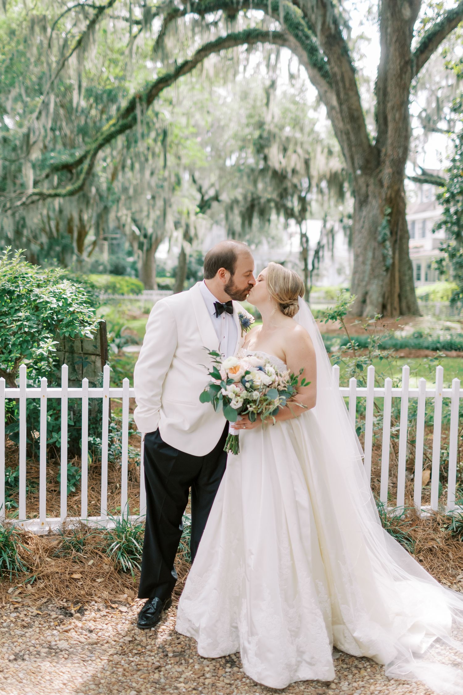 A romantic wedding couple embraces under Spanish moss-draped trees behind a white picket fence on Bluff Drive in Isle of Hope.