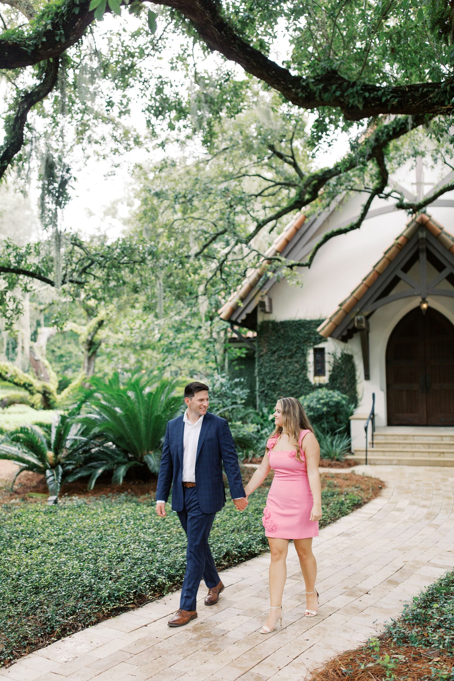 Well-dressed couple walking hand in hand along a pathway near a white stone chapel with Spanish moss draped oak trees in Sea Island, GA