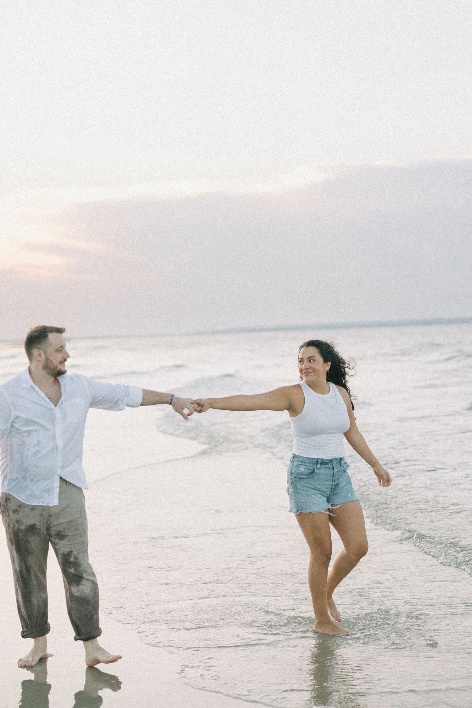 A couple holds hands while walking along a serene beach at sunset with waves gently lapping at their feet.