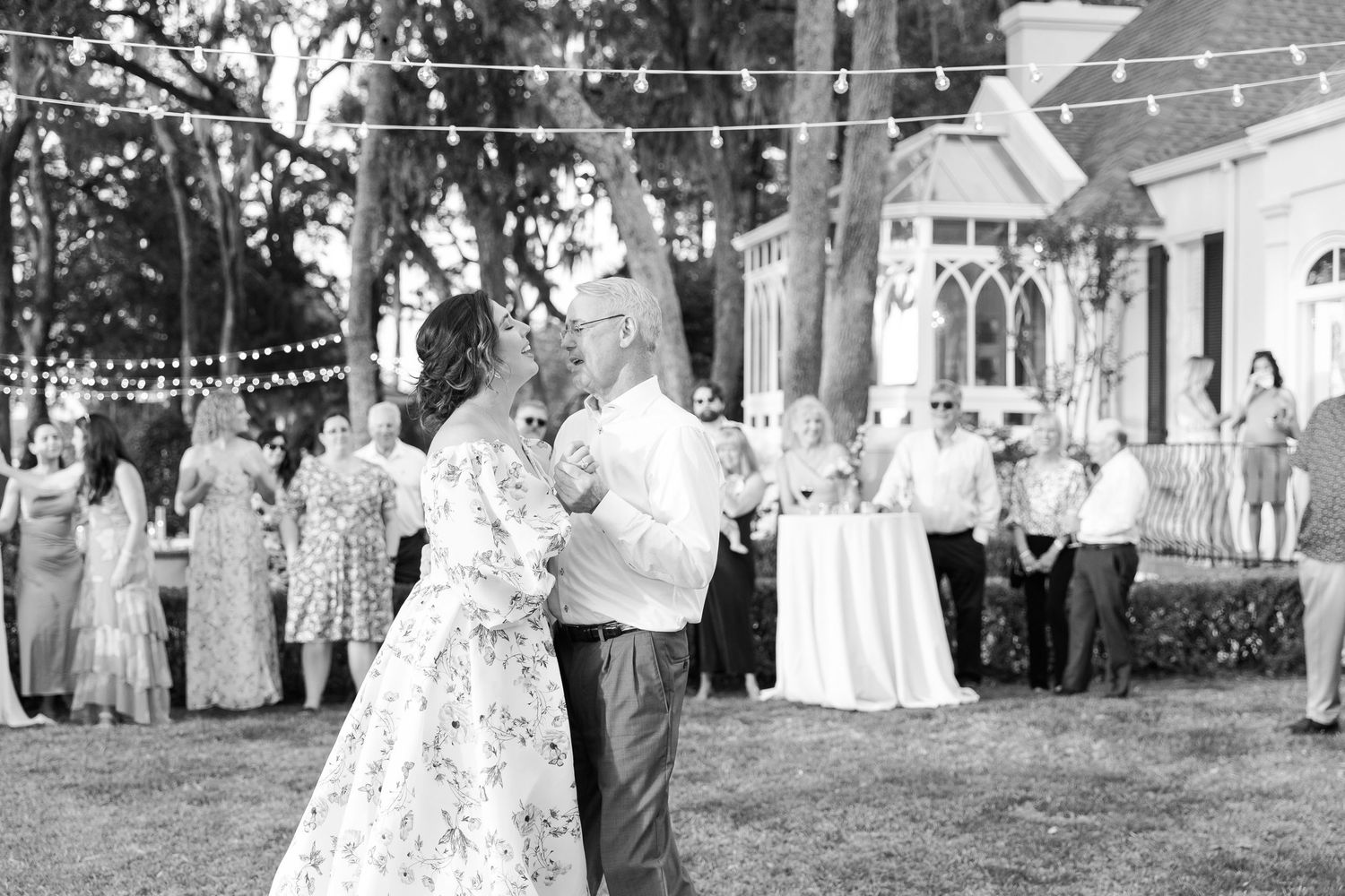 A celebration taking place outdoors with string lights and guests gathered under large oak trees.
