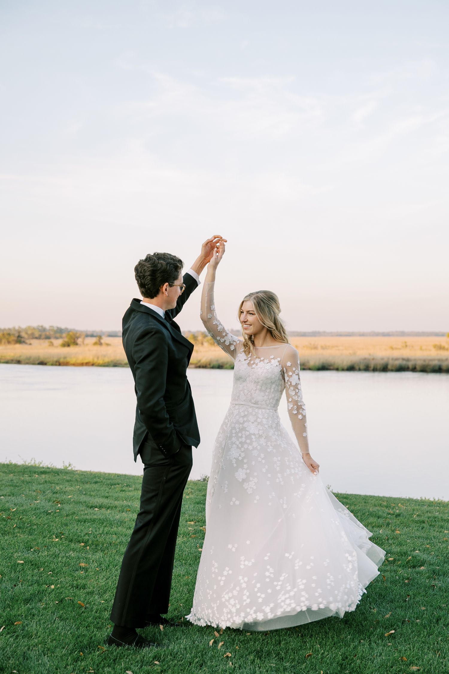 A couple in wedding attire dances together on a grassy lawn beside a serene lake at sunset.