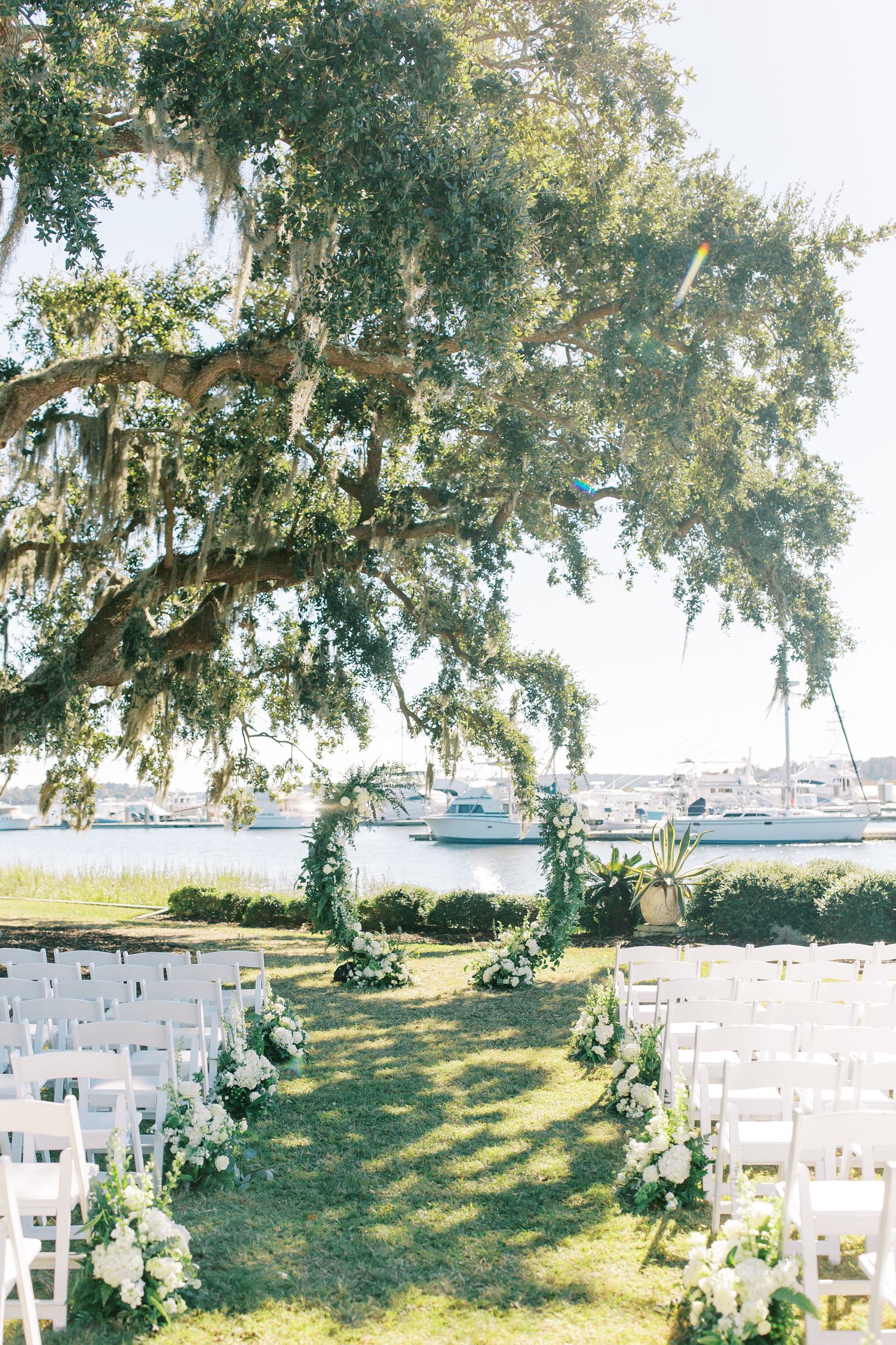 Waterfront wedding ceremony setup with white chairs and floral arrangements under a Spanish moss-draped tree.