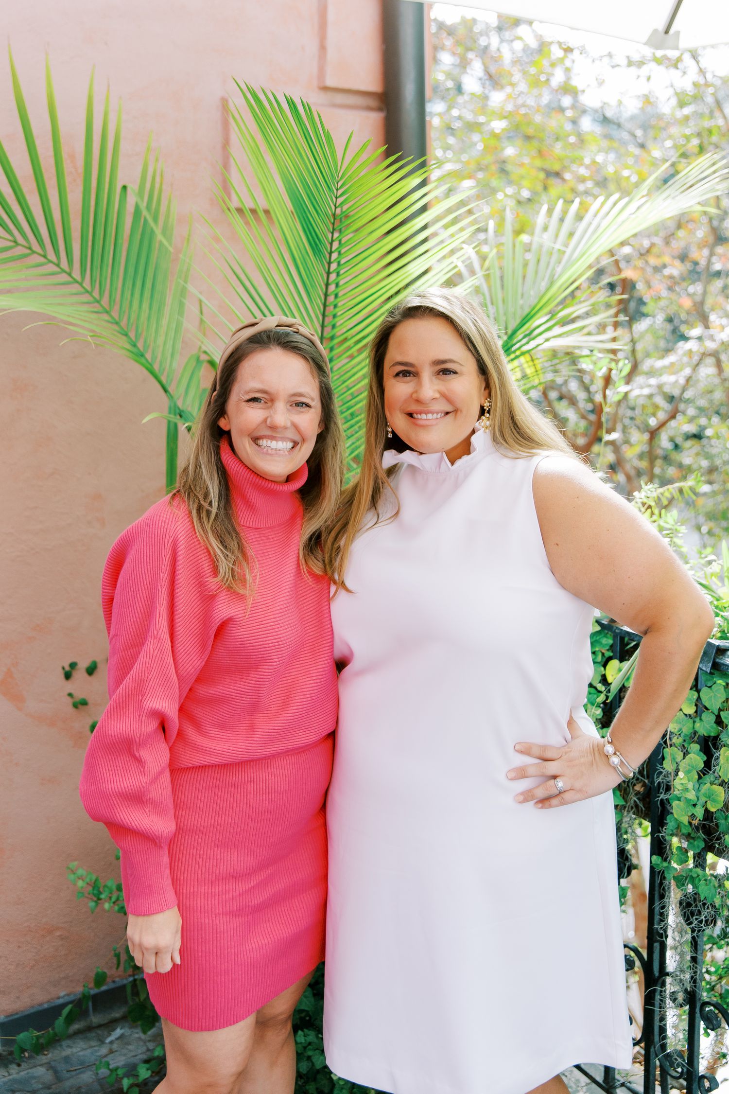 Two women posing outdoors in bright dresses against a pink wall with tropical palm fronds.