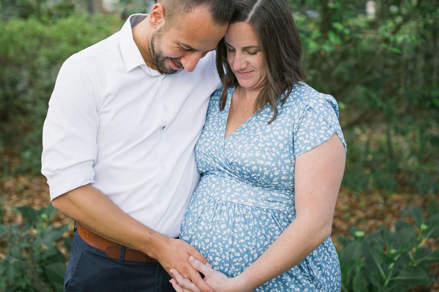 Couple wearing coordinating white and blue outfits embrace while expecting a baby in an outdoor garden setting.