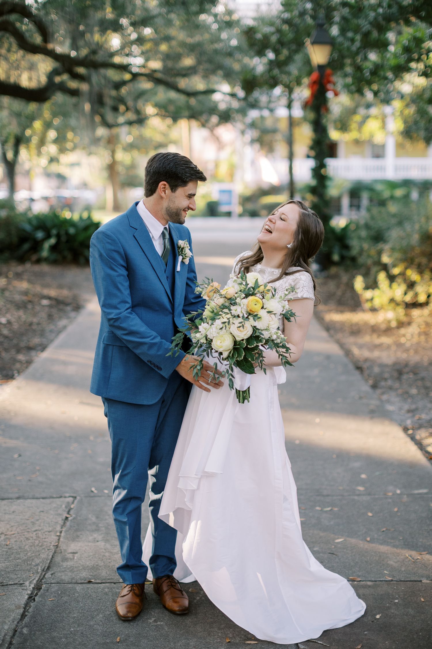 Couple shares joyful moments during outdoor wedding portraits on a tree-lined pathway.