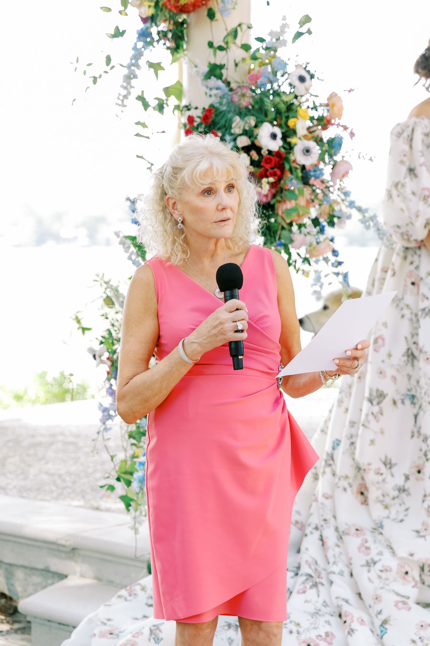 Person in coral dress giving a speech outdoors surrounded by floral arrangements.