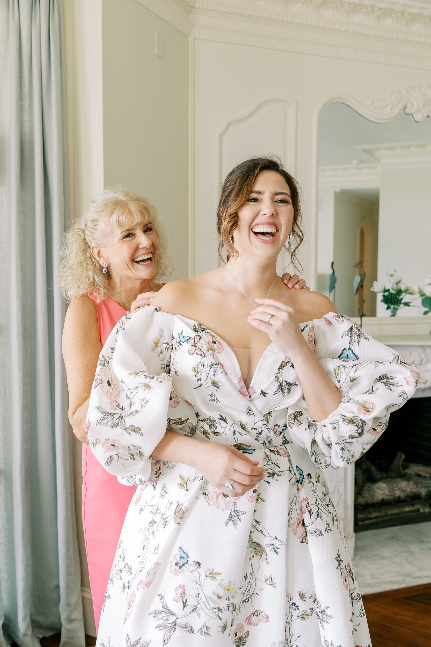 Bridesmaids in matching white floral dresses laugh joyfully together in an elegant indoor setting.