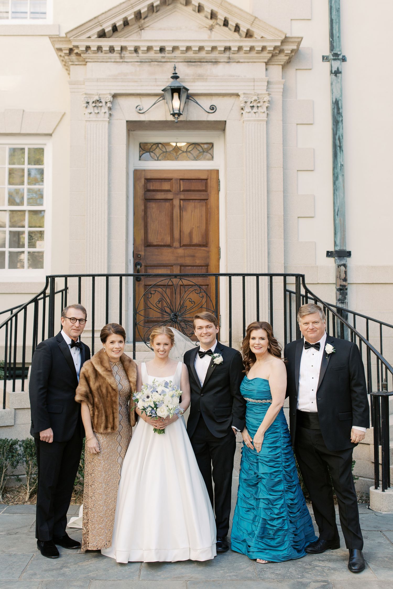 Wedding party in formal attire gathered on steps of classic white building.