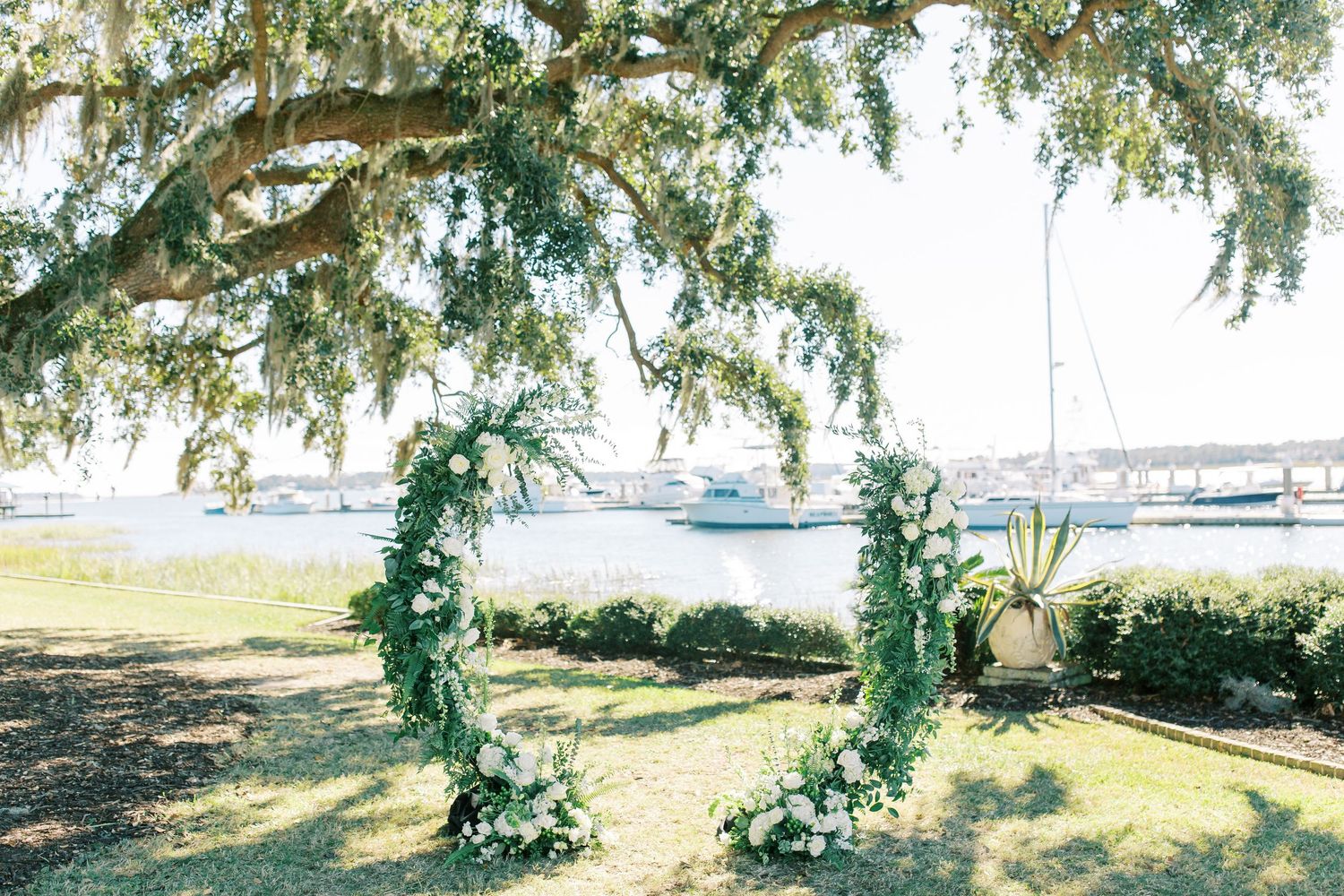 A floral wedding arch frames a waterfront view with boats and Spanish moss hanging from trees.