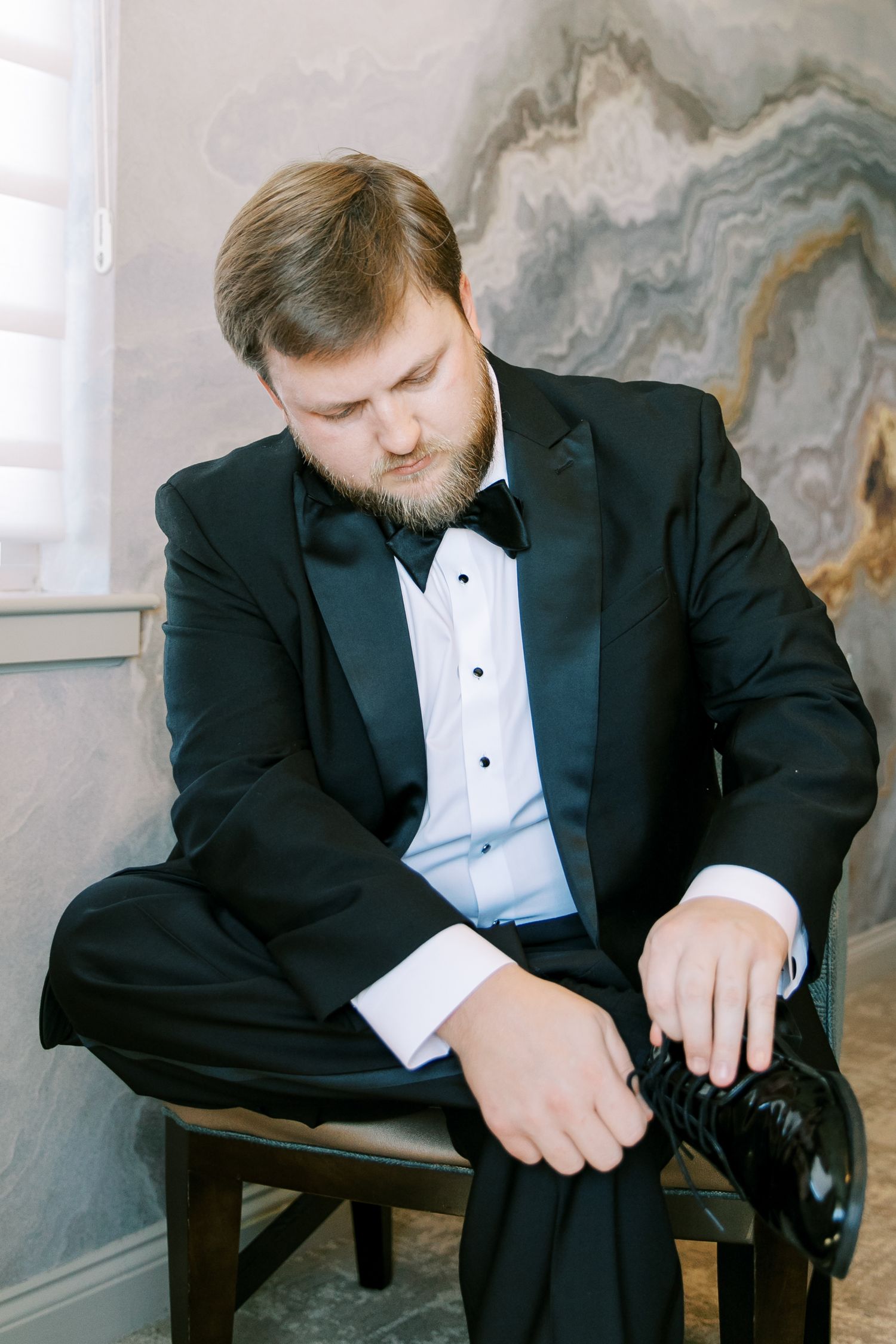 Someone in a black tuxedo adjusts their formal shoe while seated against a marbled wall background.