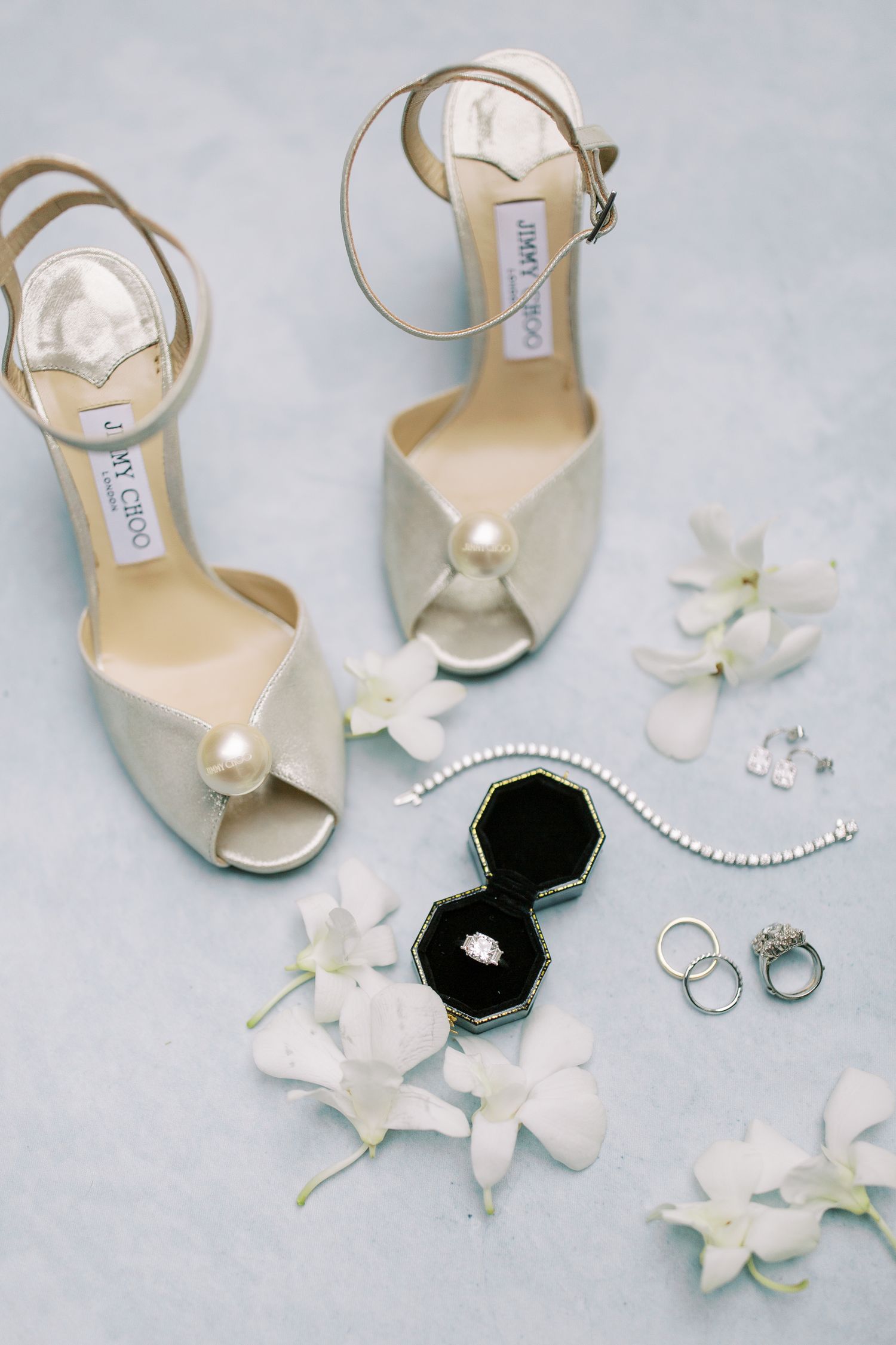 Elegant Jimmy Choo wedding shoes with pearl details alongside white orchids, jewelry, and a black velvet ring box.