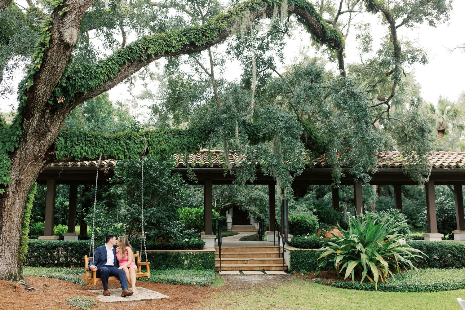 Green garden pavilion with wooden benches surrounded by oak trees draped in Spanish moss and tropical plants in Sea Island, GA