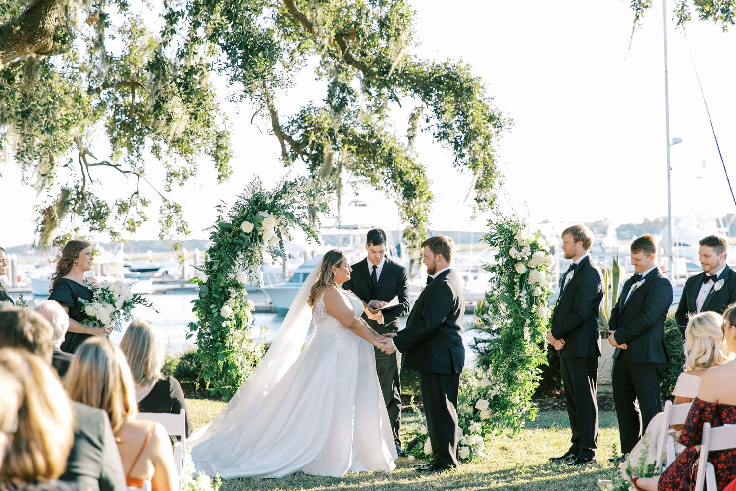 Waterfront wedding ceremony under floral arch with bridal party in formal attire.