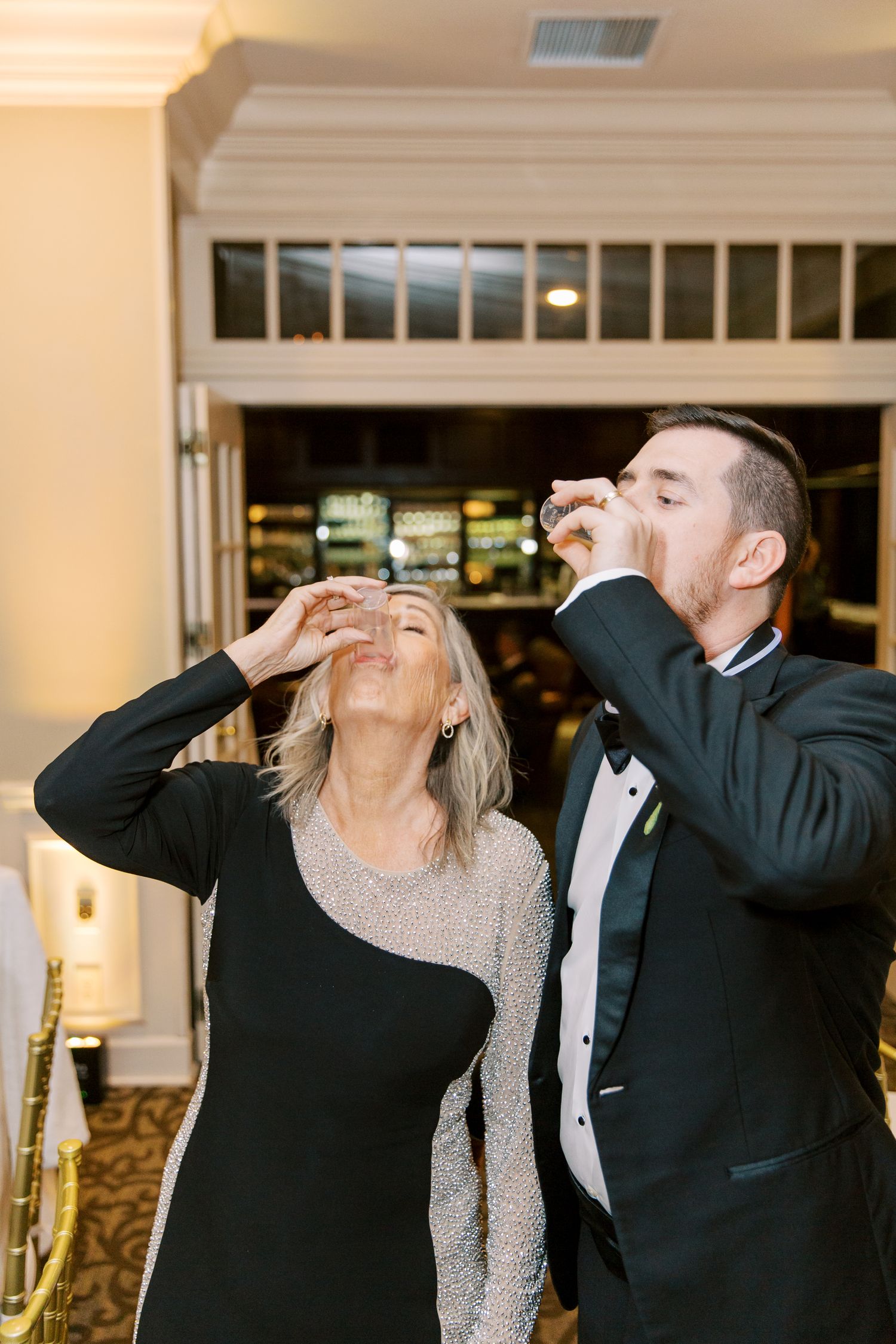A couple in formal attire taking celebratory shots together at their wedding reception.