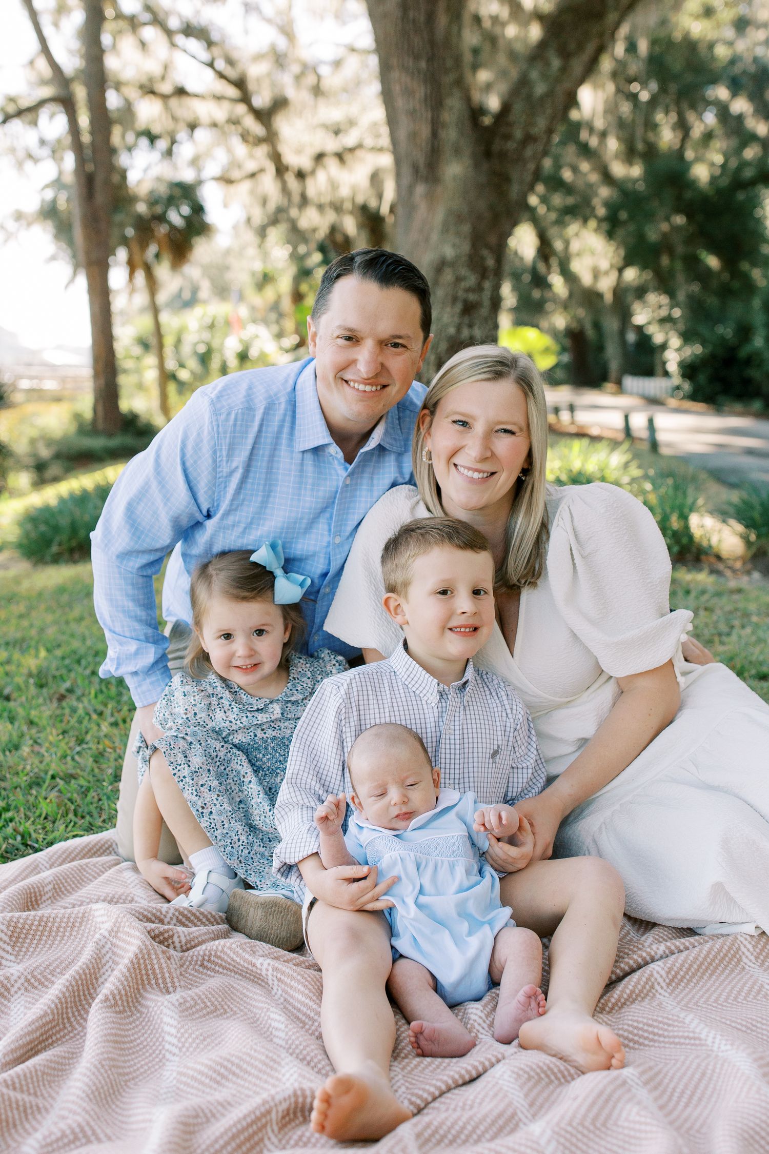 A family sits together on a blanket outdoors for a casual portrait session in a park setting.