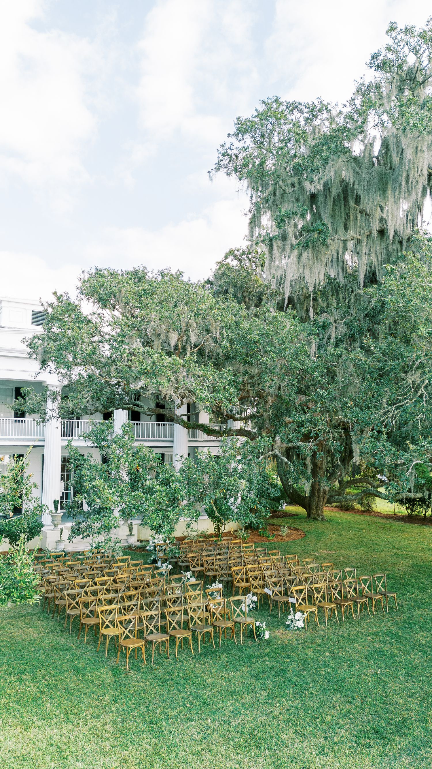 Outdoor wedding ceremony setup with wooden cross-back chairs under Spanish moss-draped trees and a white plantation house.