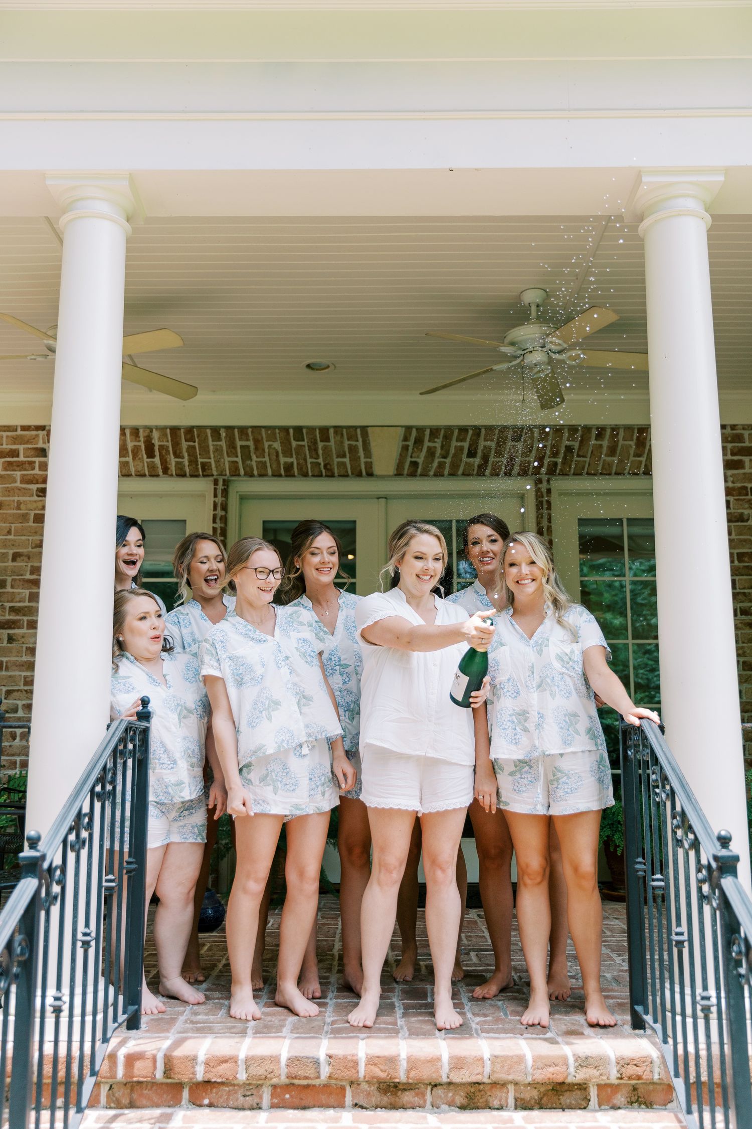 Wedding party in matching pajama sets celebrates on southern porch with champagne.