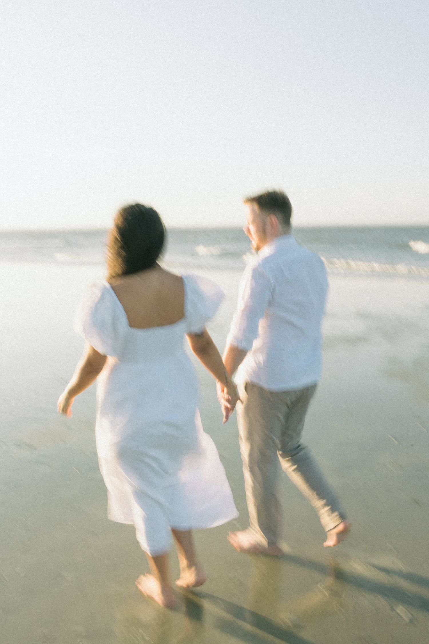 A couple in white clothing runs playfully along the beach at sunset.