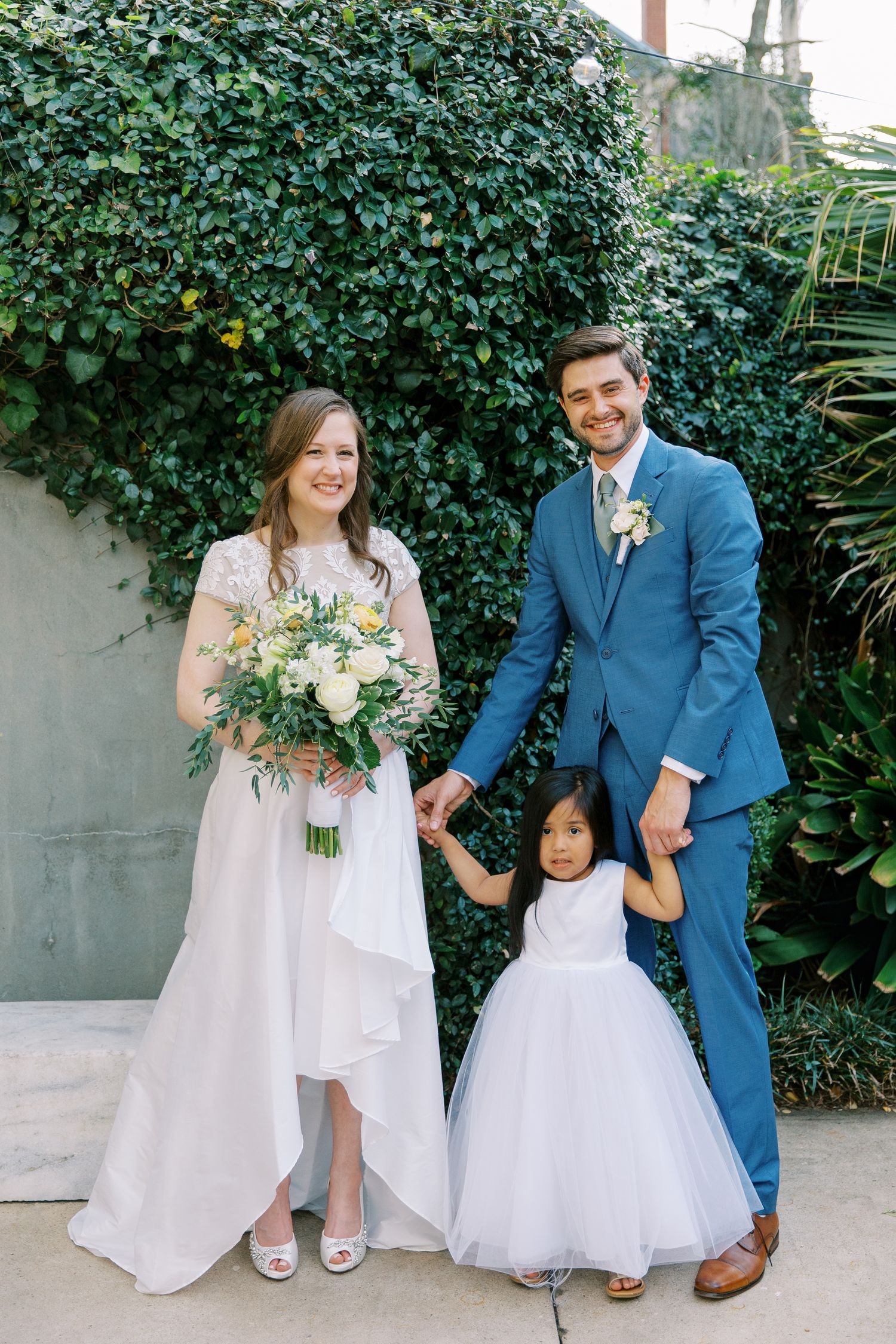 Wedding portrait against lush green ivy wall with couple and flower girl in formal attire.