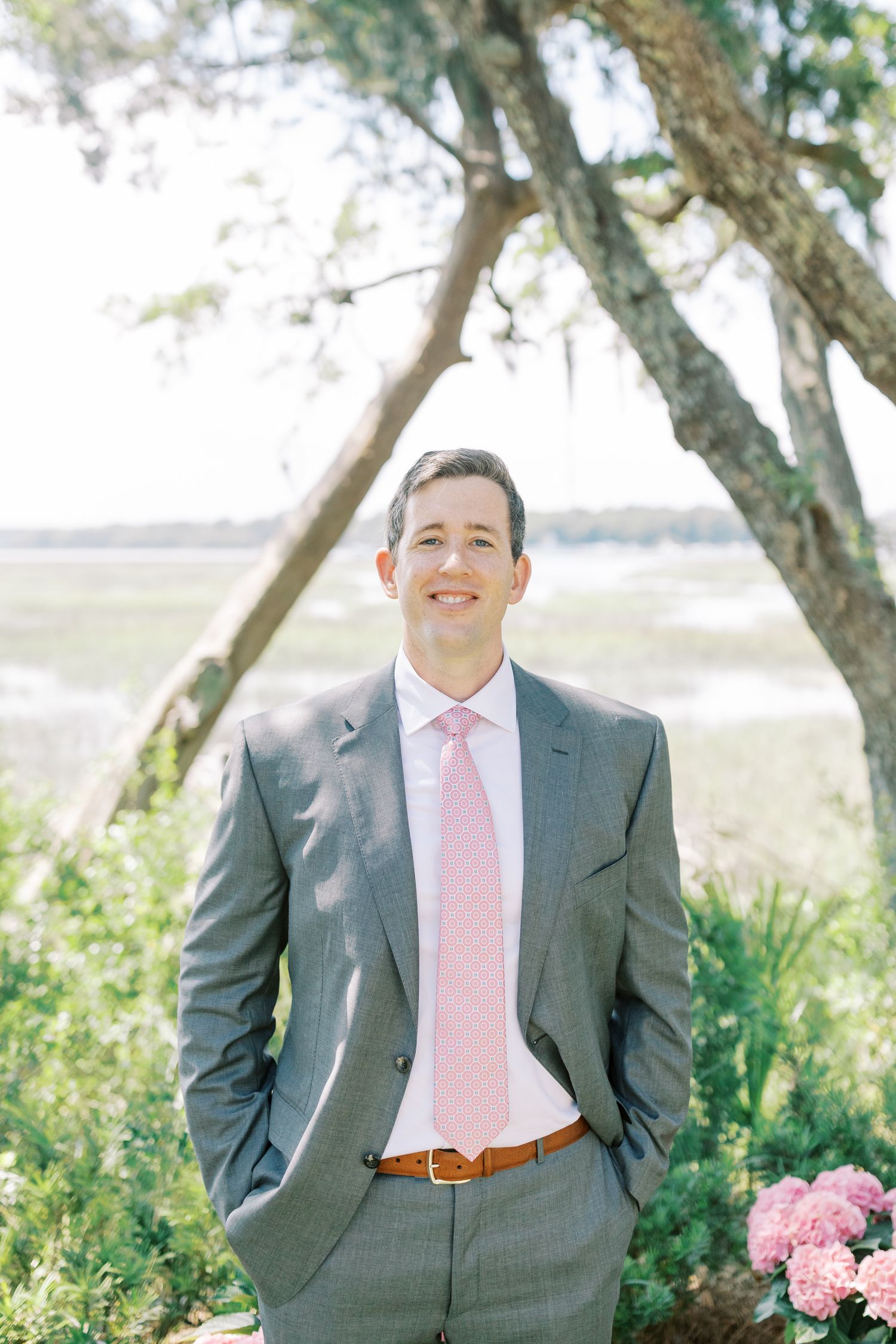 A man in grey suit and pink tie poses outdoors under trees with sunlit natural scenery in background.