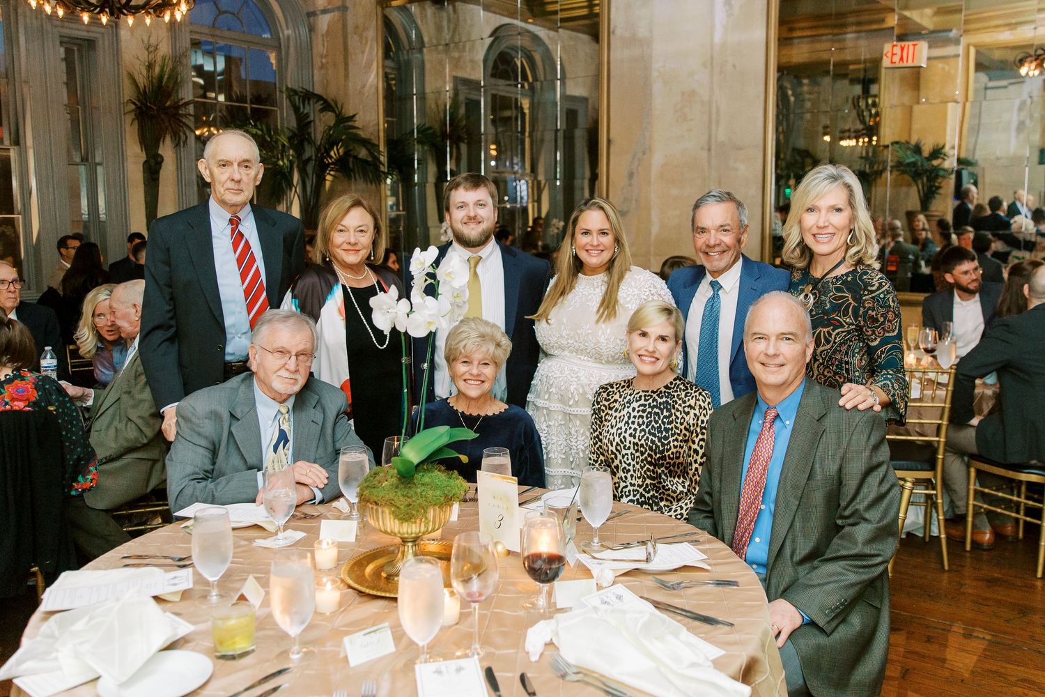 A group of formally dressed guests enjoy a meal at an elegant dining event with festive string lights overhead.