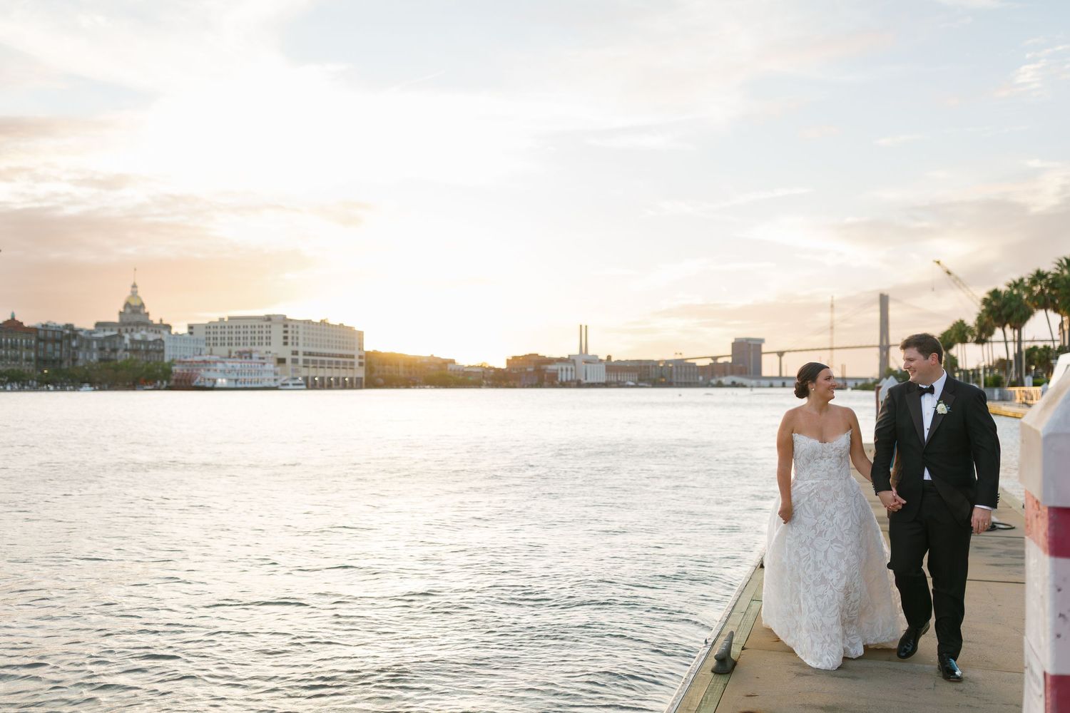 Newlyweds walk along a waterfront pier at sunset with Charleston's historic harbor skyline in the background.