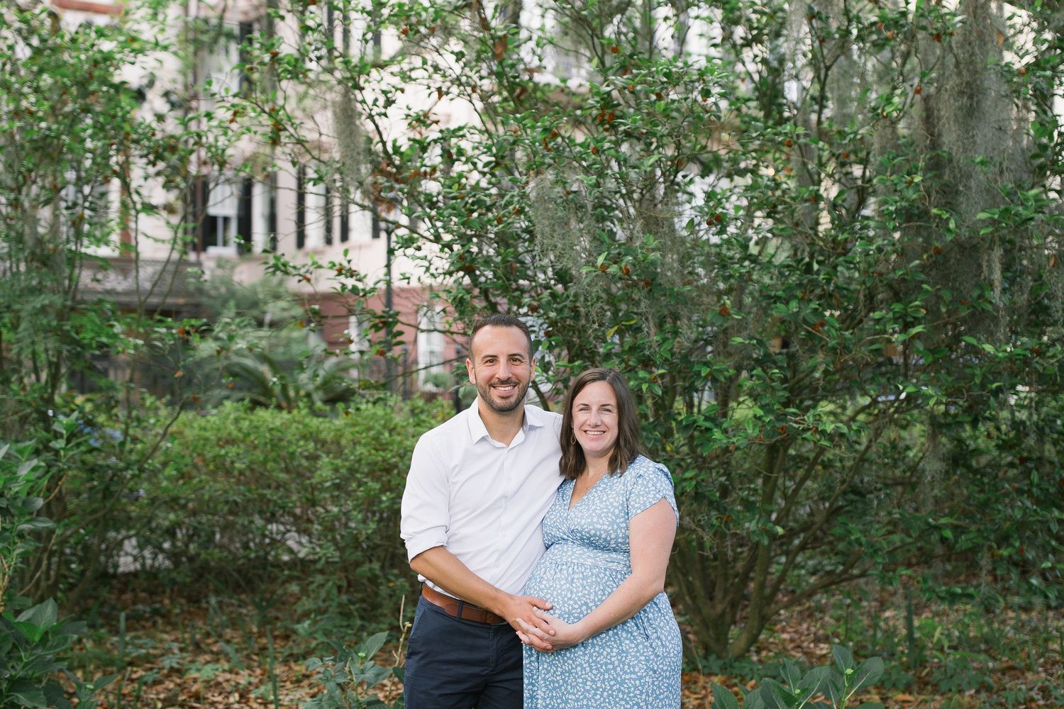 A couple stands together in a garden setting with green foliage and a house visible in the background.
