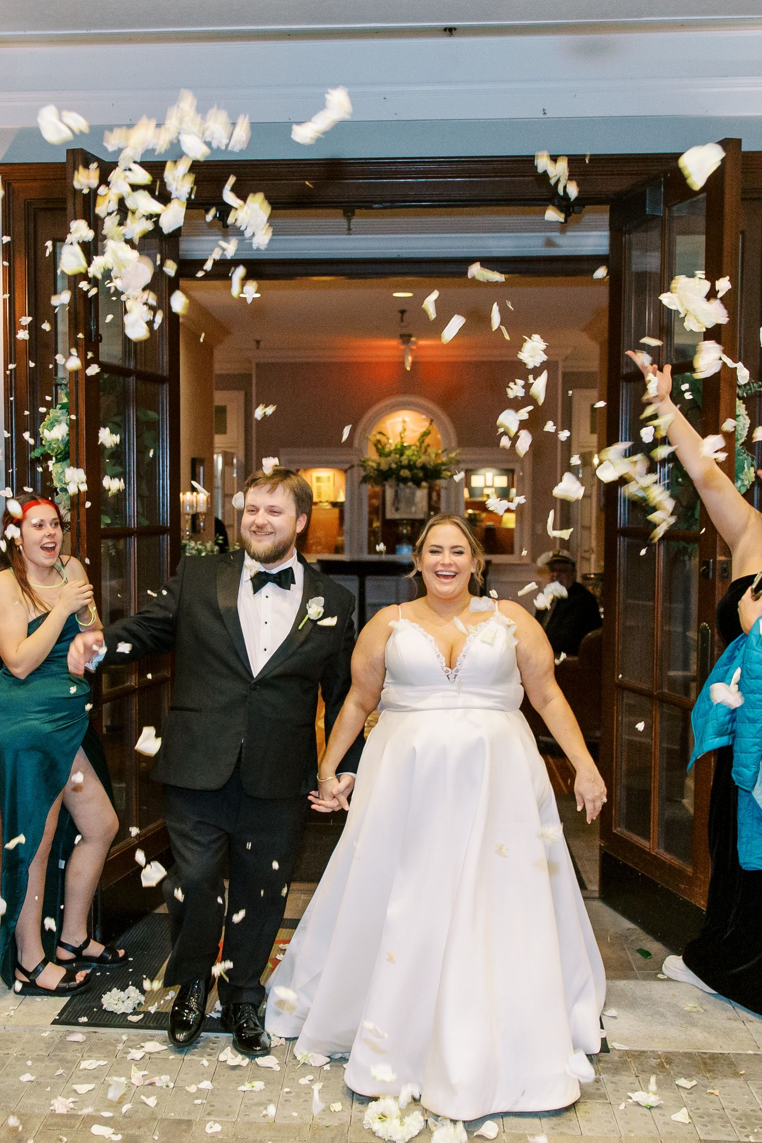 A newlywed couple exits through shower of white flower petals while guests celebrate their special moment.